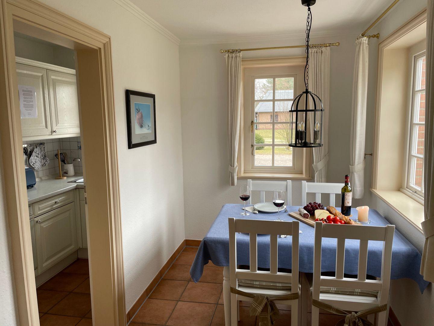 Esszimmer mit Tisch, Stühlen und Fenstern. Küche ist im Hintergrund sichtbar.