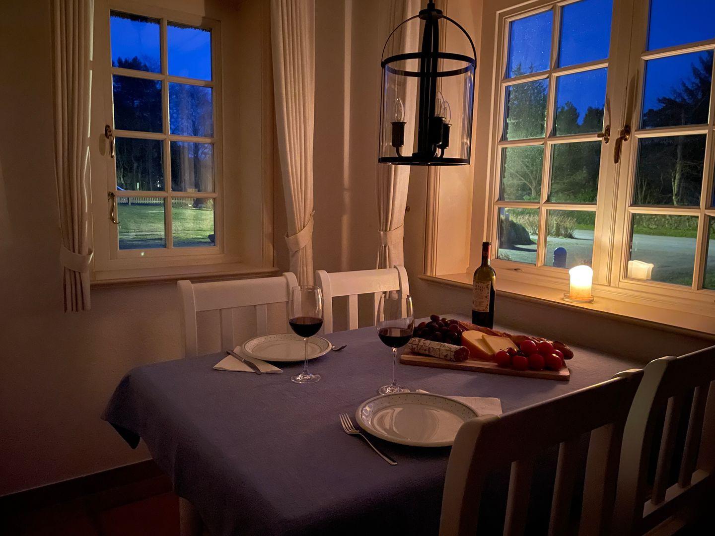 Esstisch mit Tischdecke, Gläsern, Wein und Käse vor Fenstern mit Blick auf Garten.