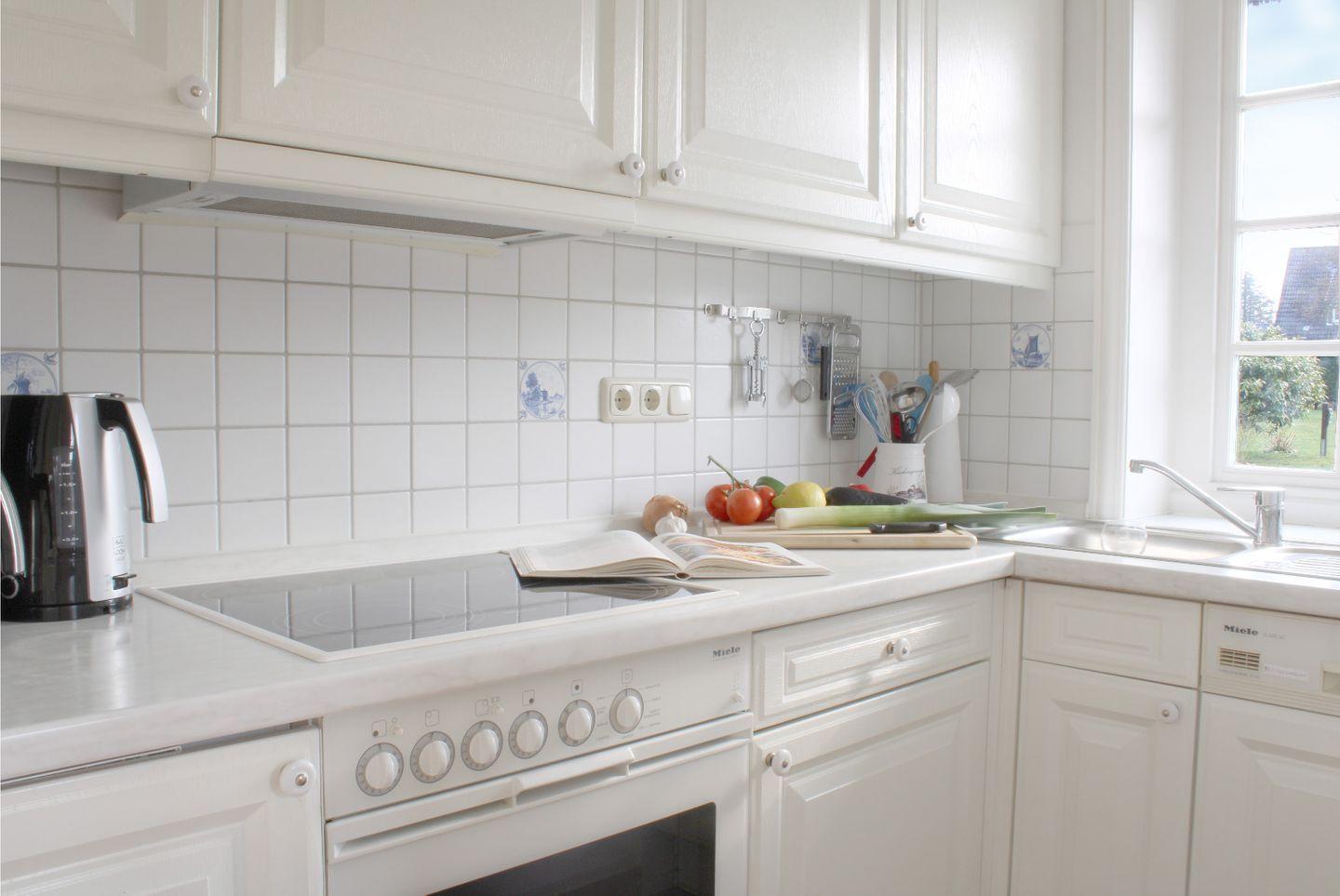Küche mit weißen Schränken, Herd und Spüle. Auf der Arbeitsfläche liegen Obst, Gemüse und ein Buch.