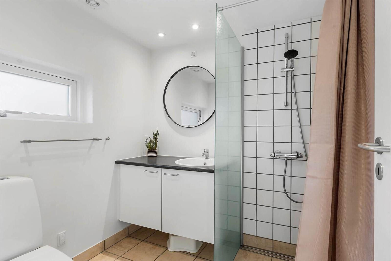 Bathroom with shower, sink and toilet. White walls, round mirror and brown tiles.