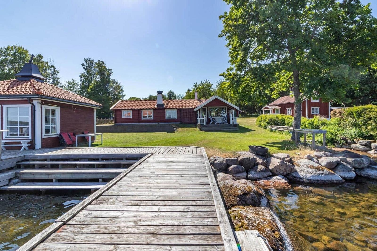 Holzsteg führt zu roten Häusern am Wasser mit Garten und Bäumen.