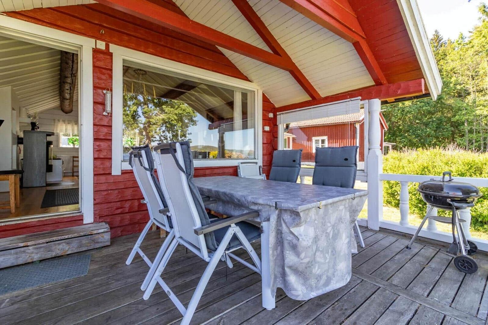 Terrasse mit Tisch, Stühlen und Grill. Roter Holzaufbau und Sicht in die Küche.