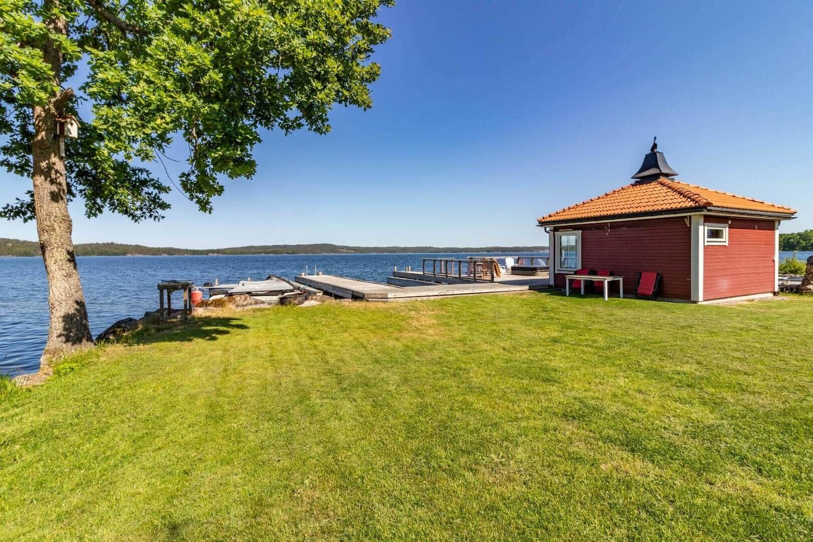 Rotes Bootshaus am See mit Terrasse und Liegeplatz. Grasfläche und Baum im Vordergrund.