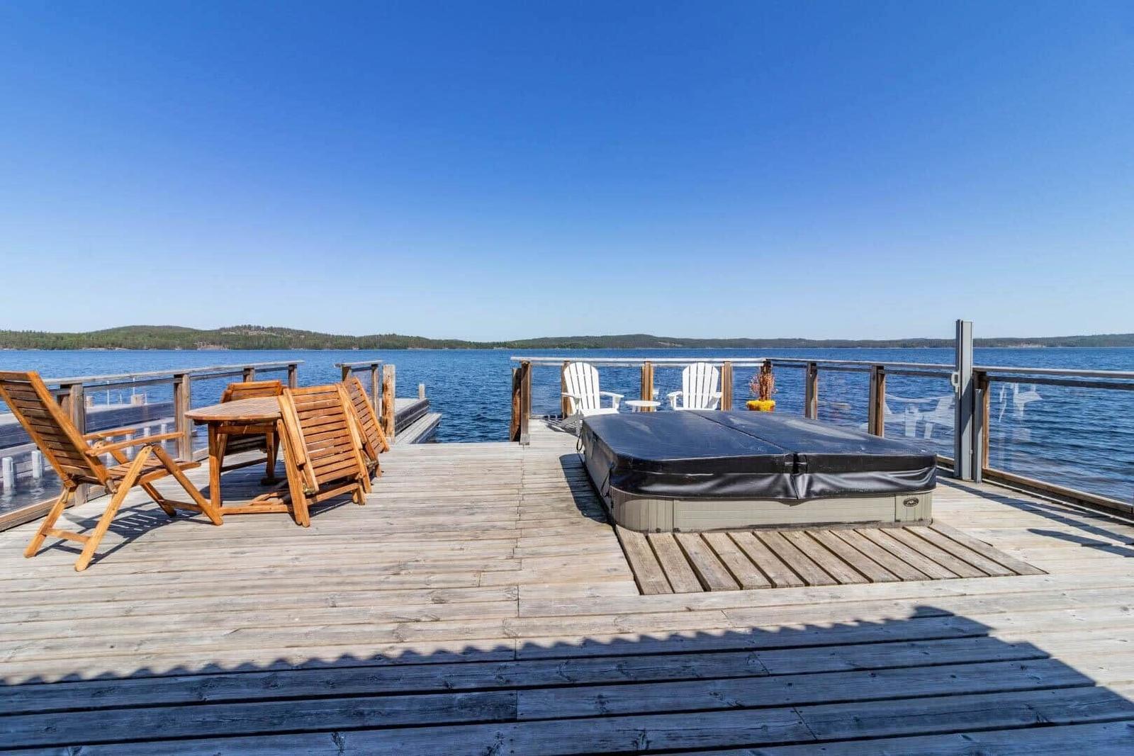 Holzterrasse mit Liegestühlen, Tisch und Whirlpool am Wasser.