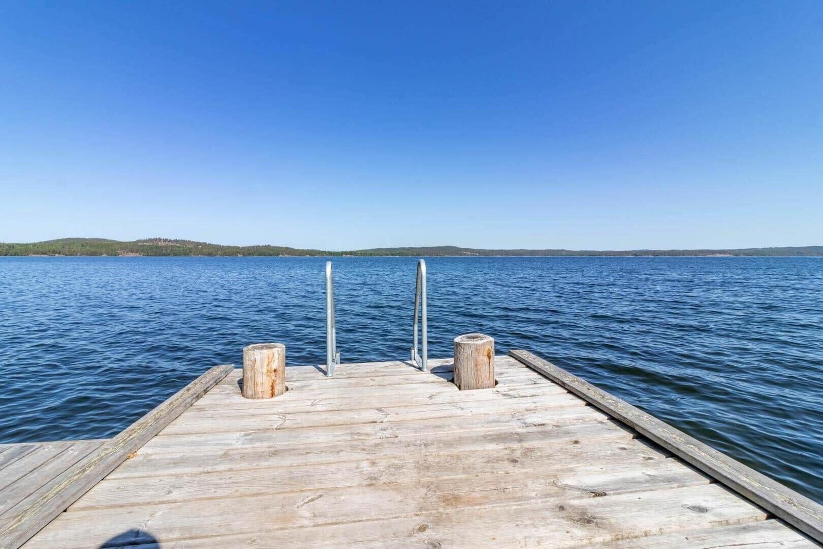 Holzsteg mit Metallgeländer führt ins Wasser. Horizont mit bewaldeten Hügeln unter blauem Himmel.