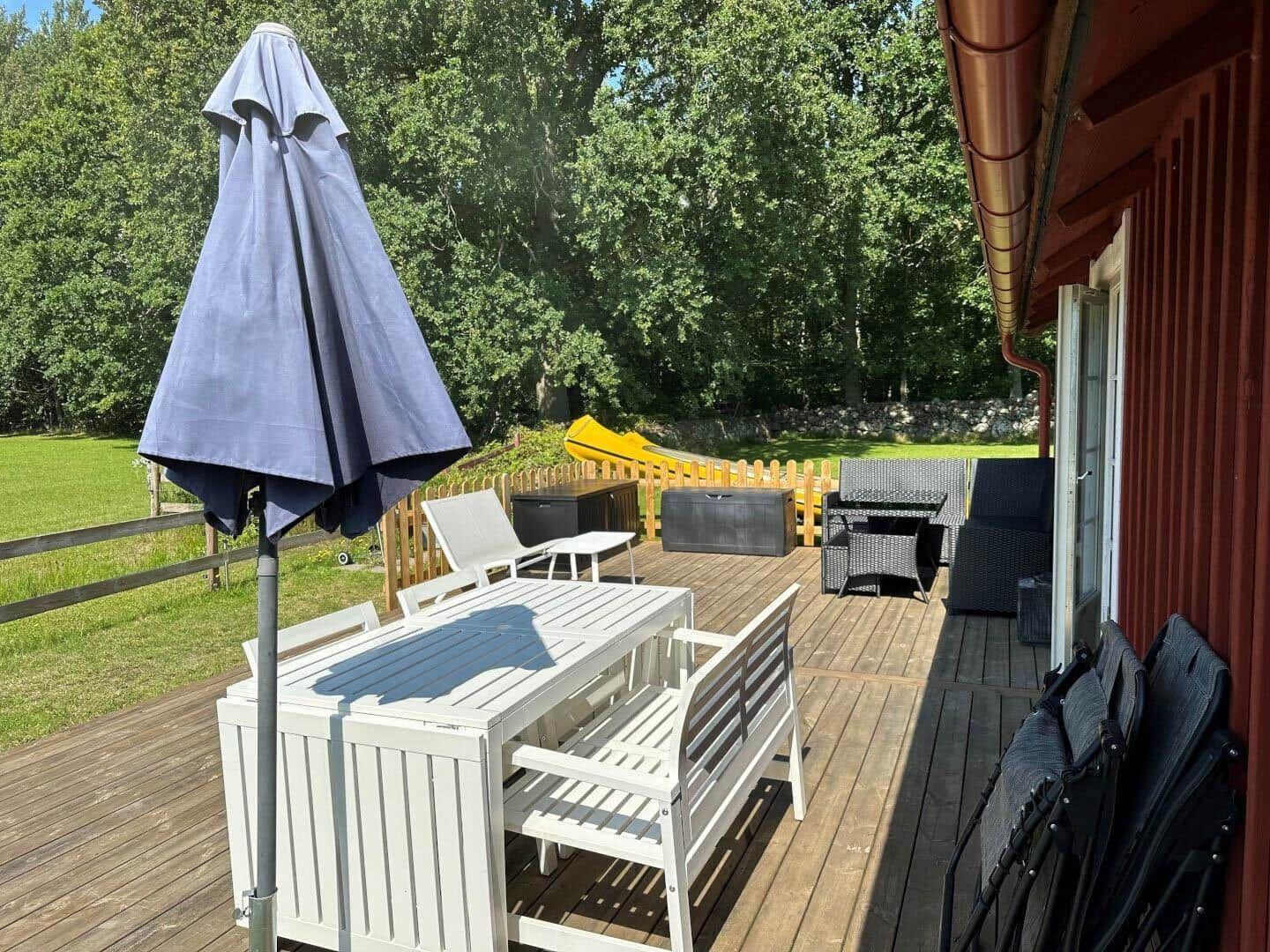 Terrasse mit weißem Tisch, Stühlen und geschlossenem Sonnenschirm. Im Hintergrund Grünfläche und Spielplatz.