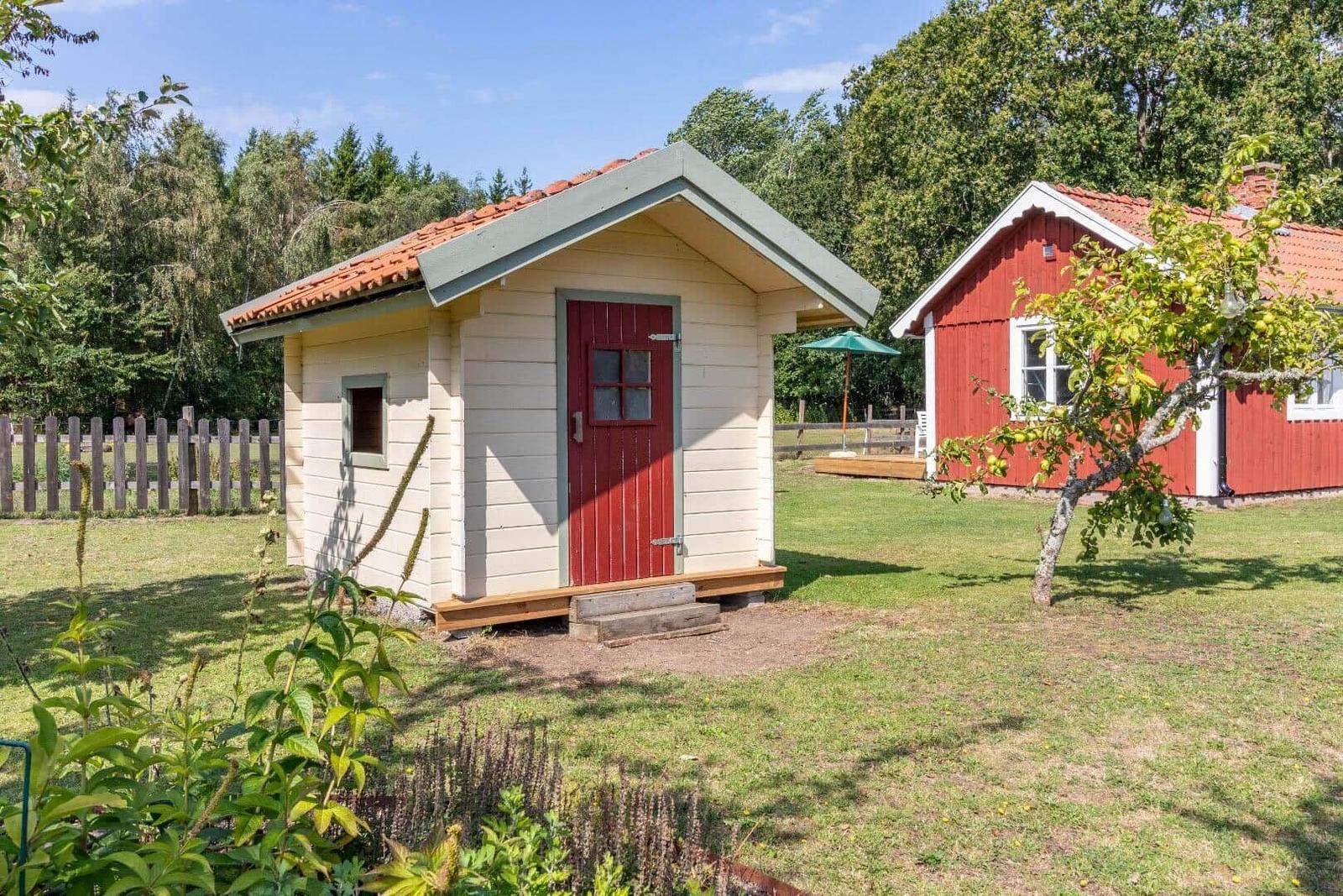Ein kleines Haus mit roter Tür und Terrakottadach steht auf einer Wiese neben einem größeren roten Haus.