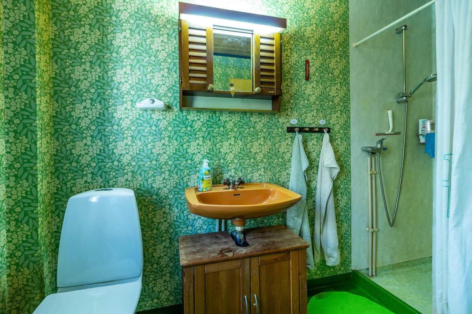 Badezimmer mit grünem Tapetenmuster, Waschbecken auf Holzschrank, Dusche und Toilette.