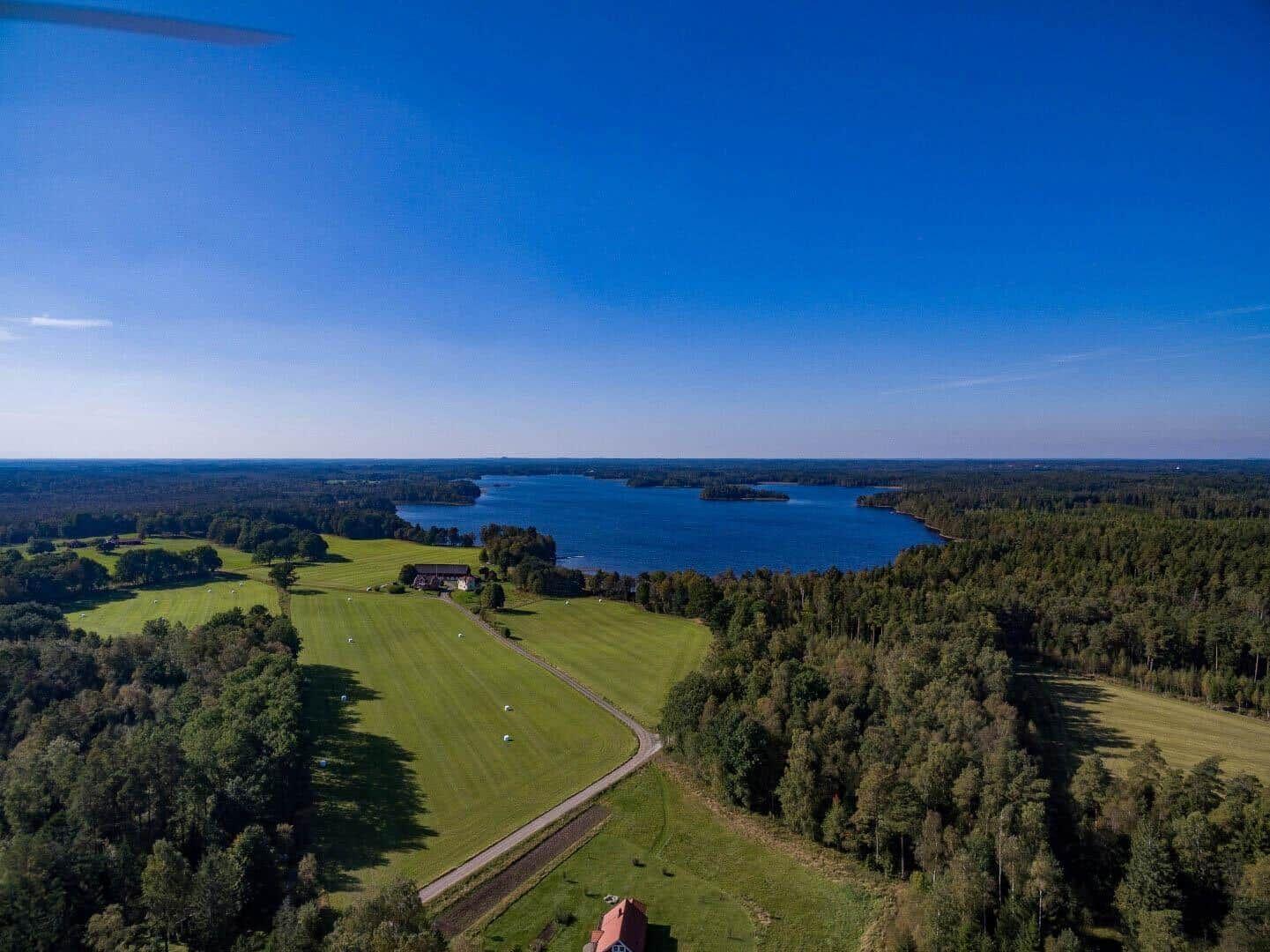 Aerial view of a lake surrounded by forests and green fields with a house nearby.