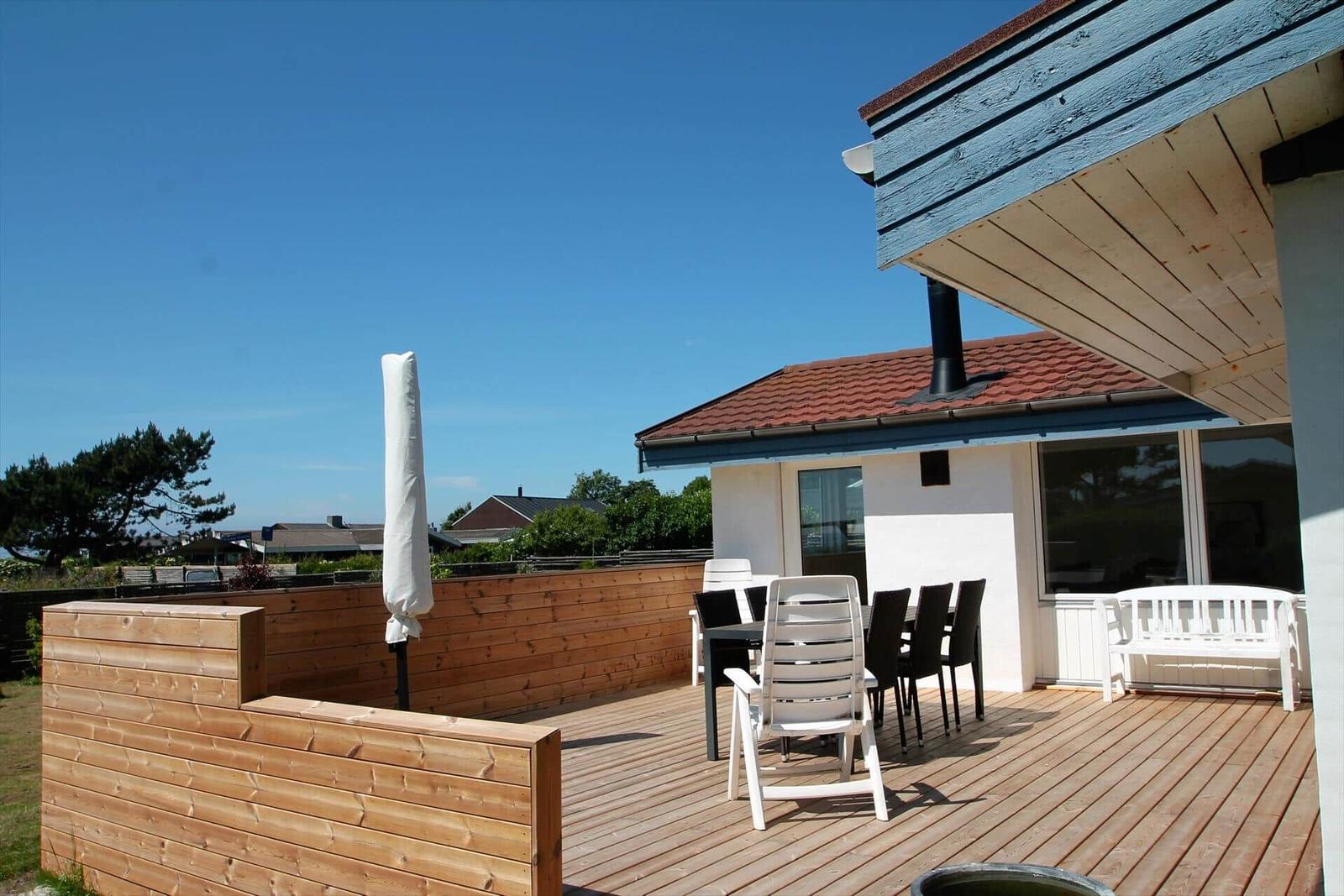 Terrasse mit Holzdecke, Tisch, Stühlen und Schutzdach. Hintergrund: Haus und Bäume.