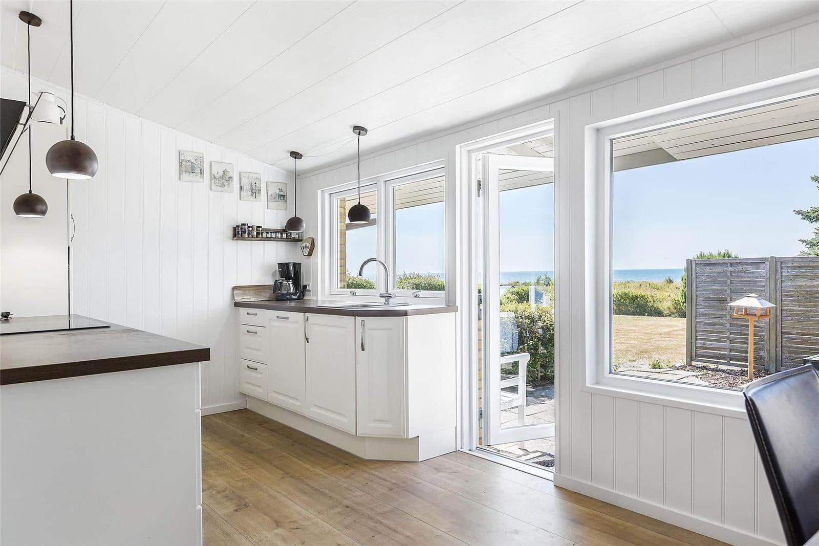 Küche mit Blick auf den Strand und Terrasse. Weiße Schränke, Holzboden und Fensterfront.