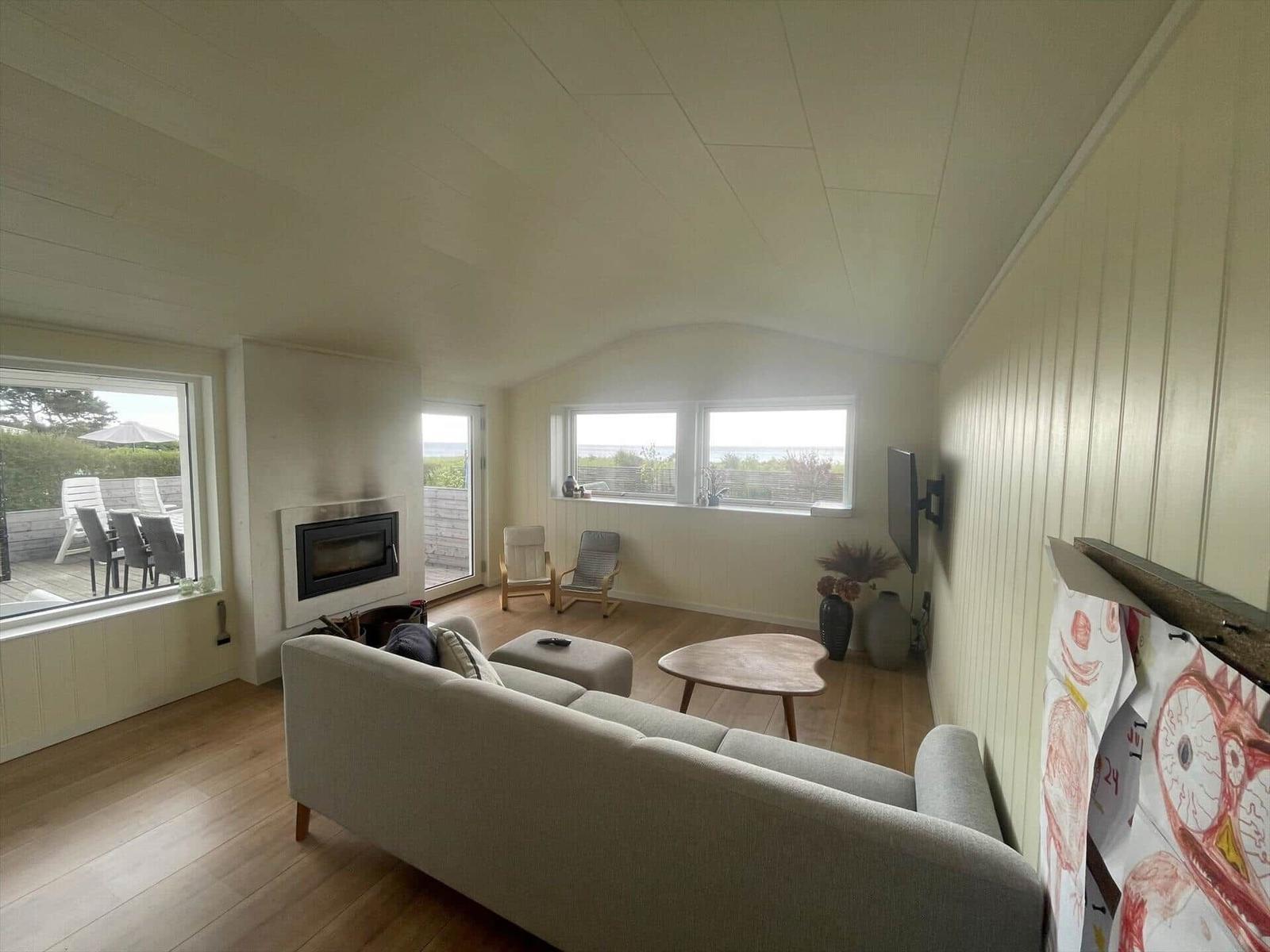 Wohnraum mit Sofa, Kamin und Fenstern zum See. Holzboden und Wandverkleidung.