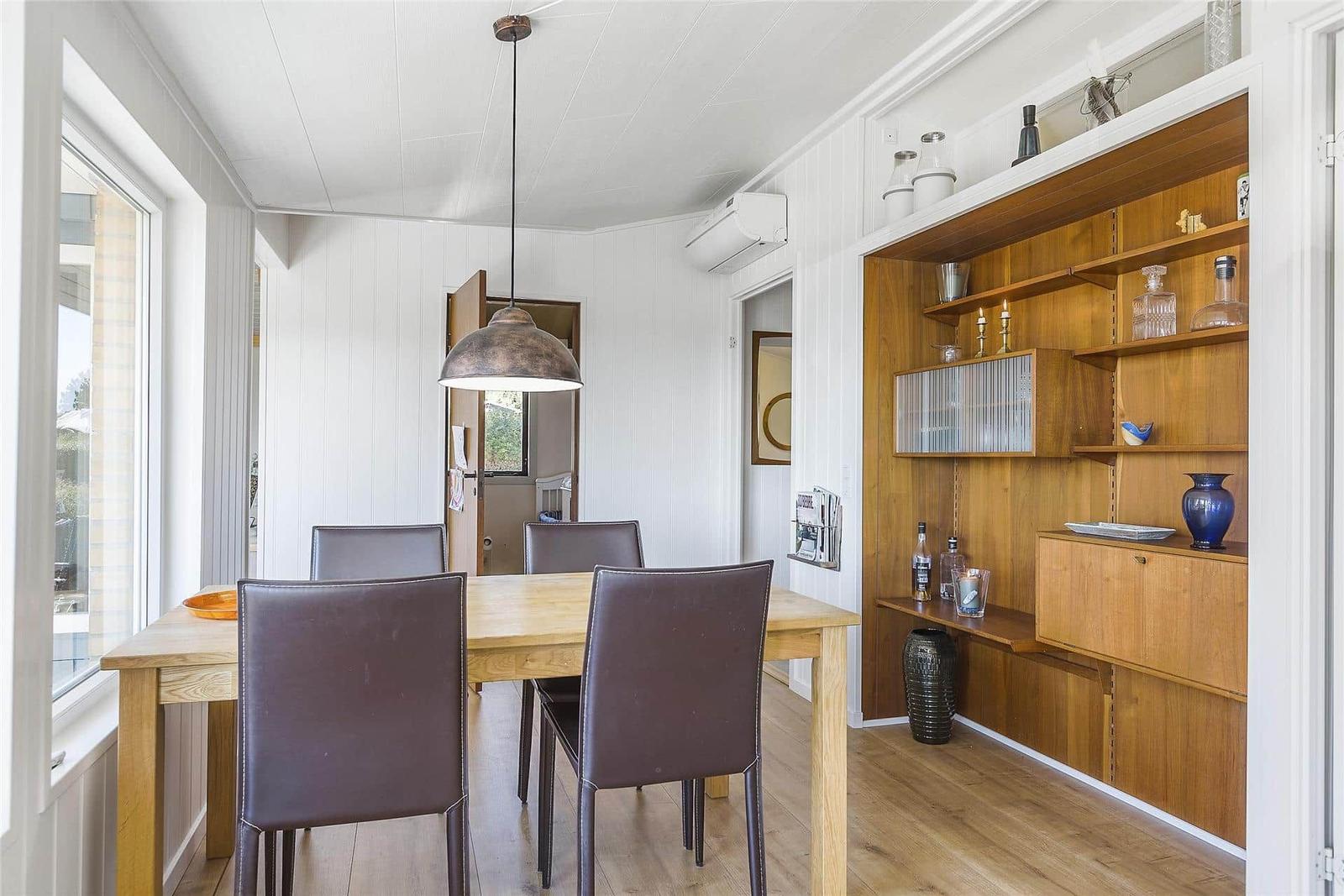 Esszimmer mit Holztisch, vier Stühlen und Holzwandregal. Deckenleuchte und Fenster sichtbar.
