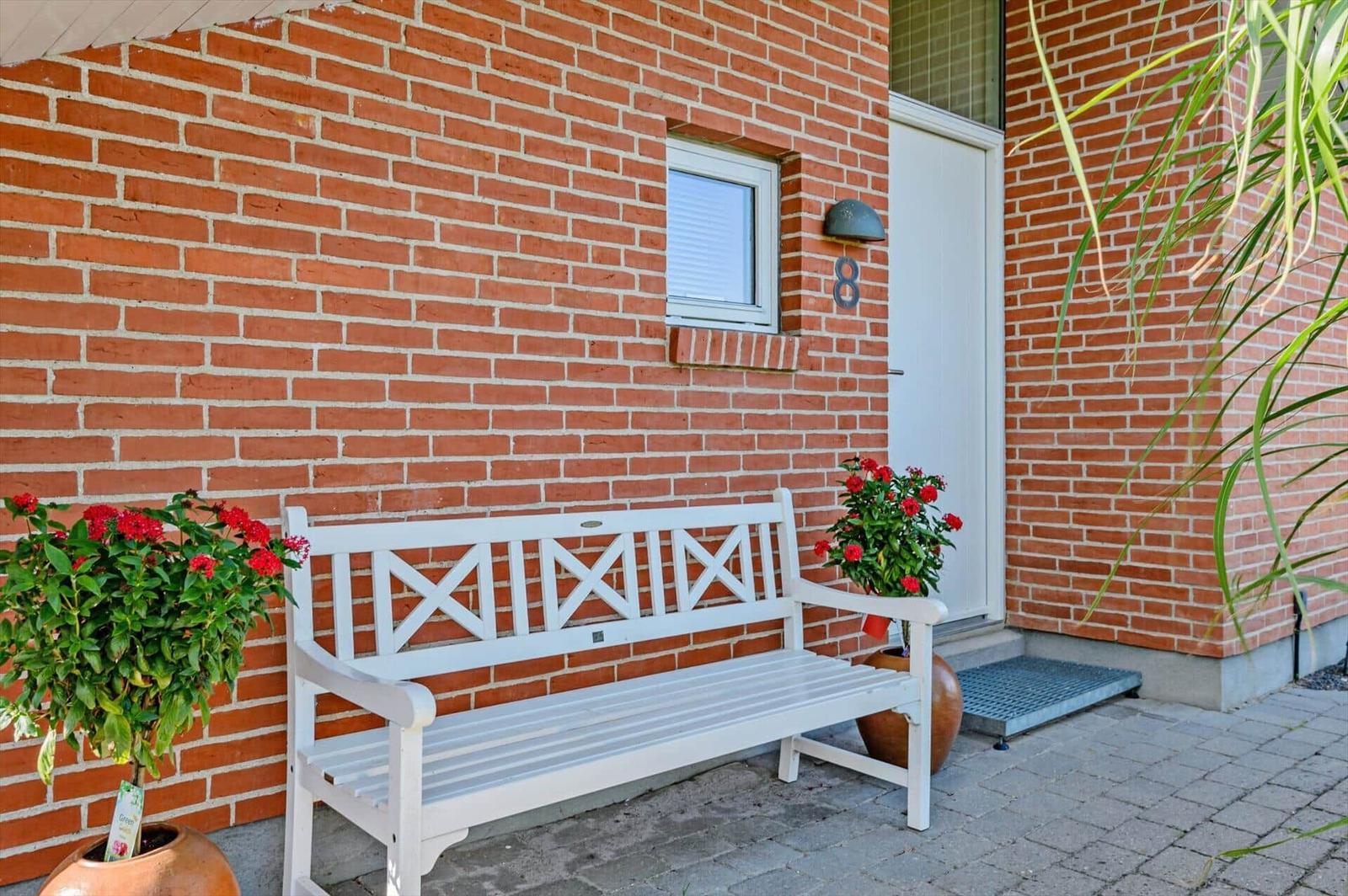 A white bench stands before a red brick wall beside a door with house number 8.