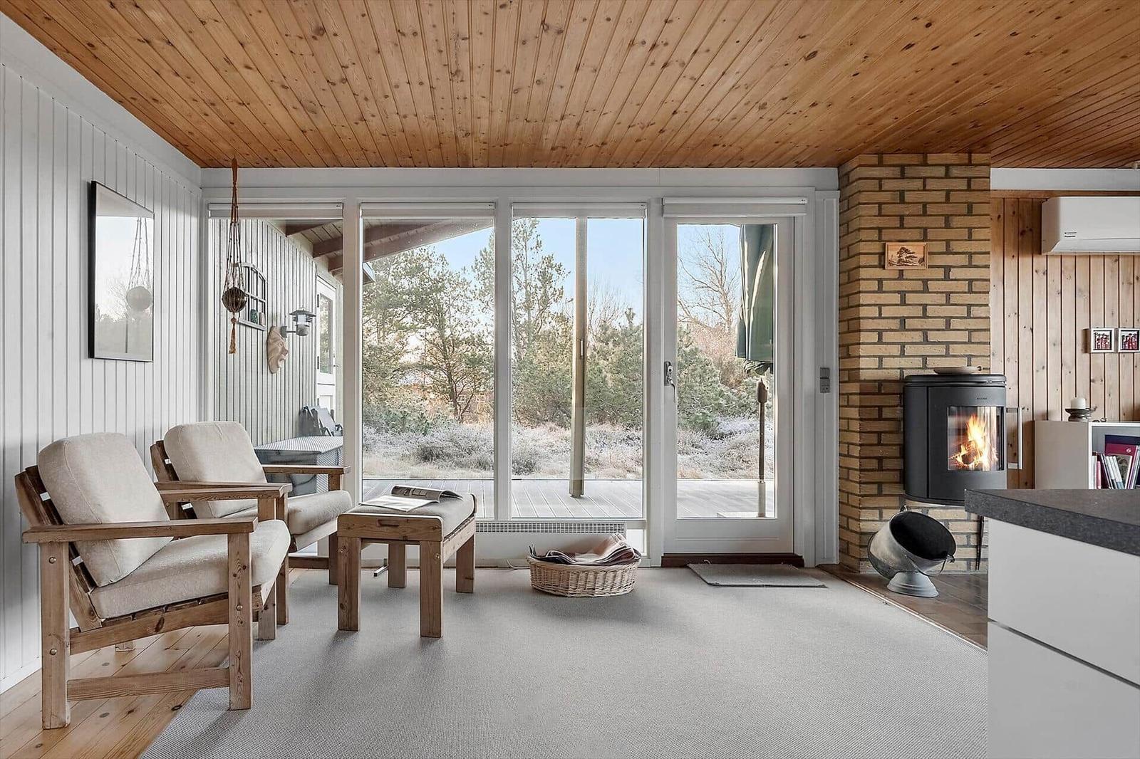 Wohnraum mit Holzdecke, Sitzgelegenheit und Kaminofen. Durchgang zur Terrasse mit Blick auf Wald.