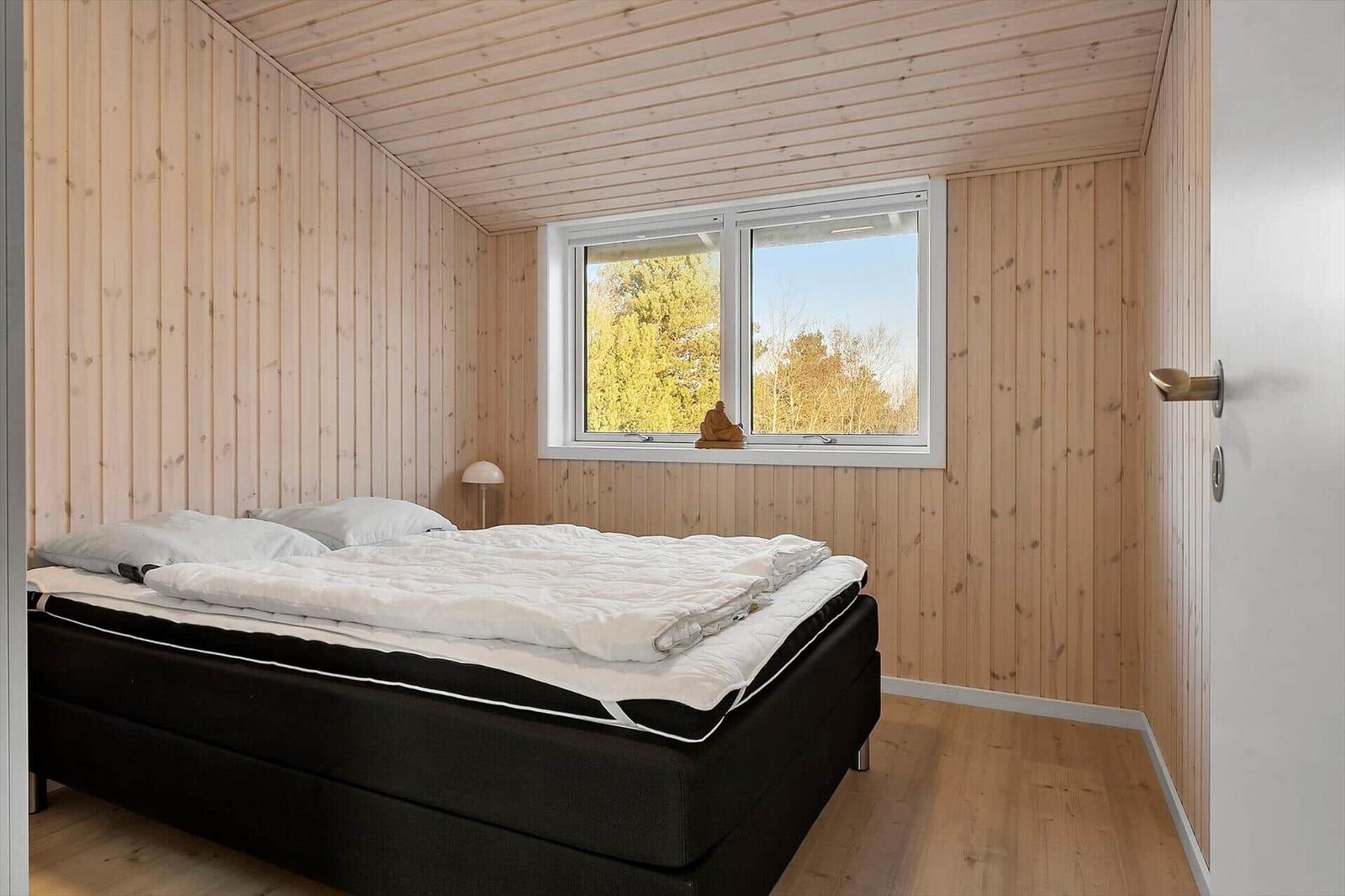 Schlafzimmer mit Holzwand, Bett und Fenster mit Blick auf Bäume.