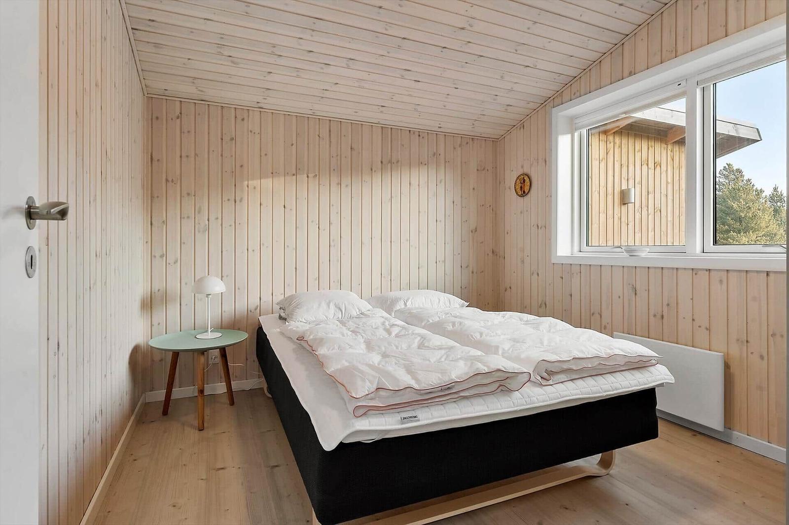 Schlafzimmer mit Holzwandverkleidung, Bett und Fenster mit Blick auf Wald.