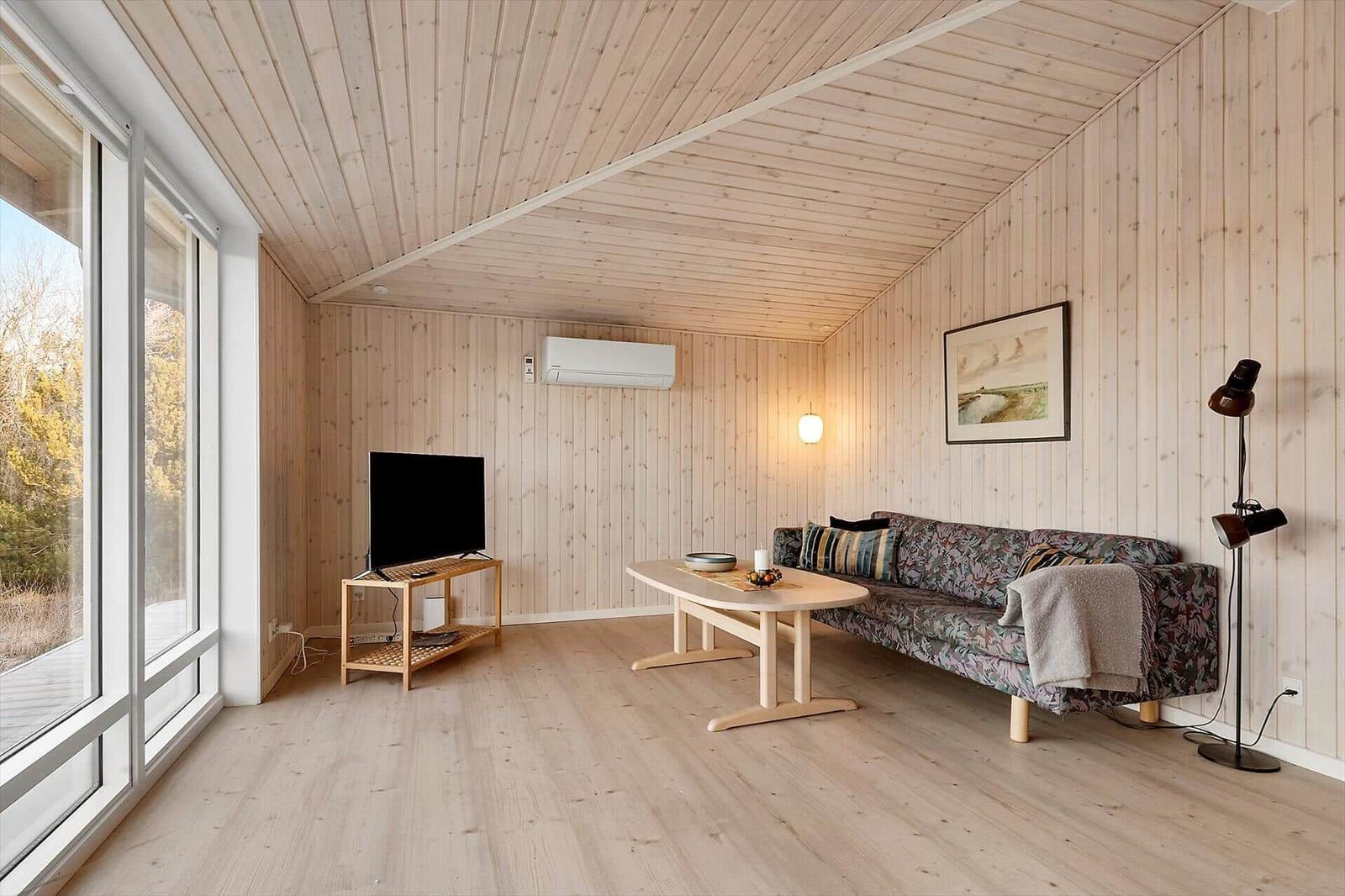 Wohnzimmer mit Holzwand, Sofa, Tisch und TV. Große Fenster mit Blick nach draußen.