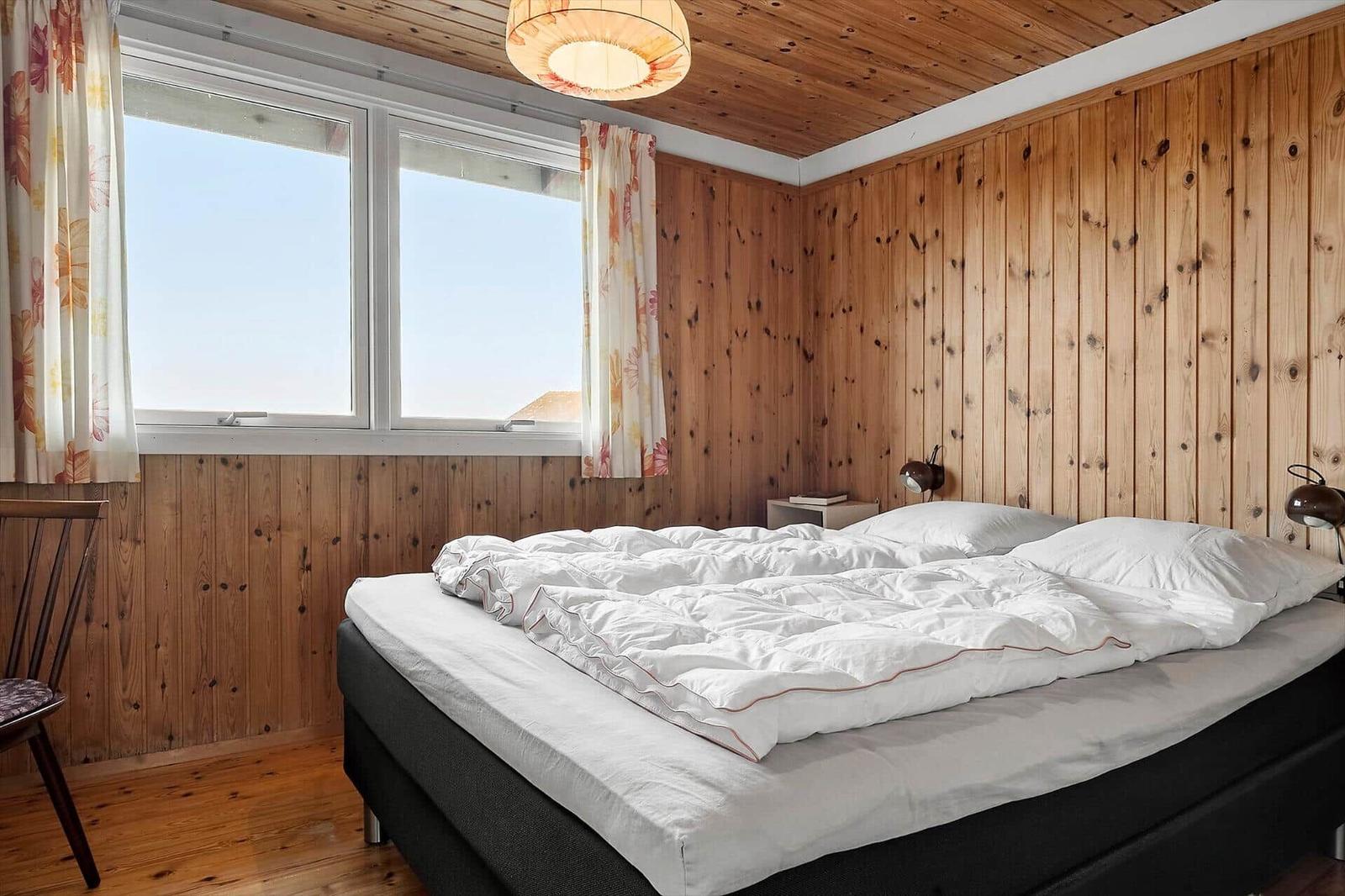 Schlafzimmer mit Holzwand, großem Fenster und Bett mit weißer Bettwäsche.