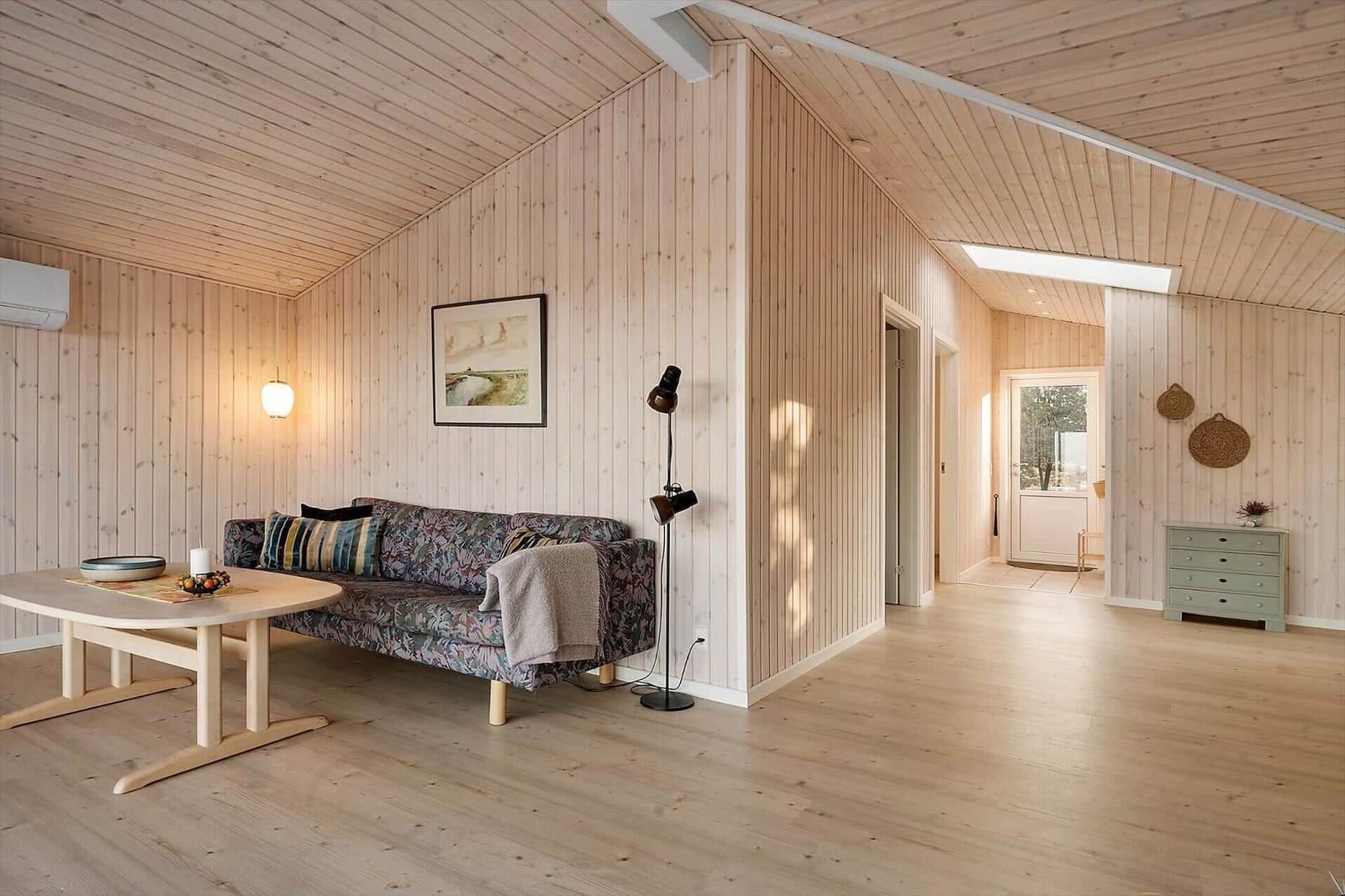 Wohnzimmer mit Holzwand, Sofa, Tisch und Stehlampe. Durchgang zu einem weiteren Raum.