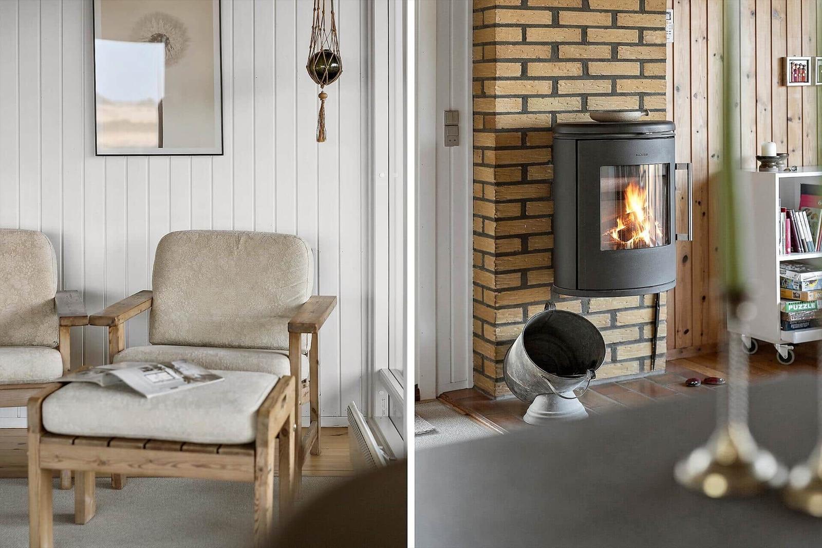 Zwei Sessel mit Holzrahmen und hellen Polstern stehen vor einer weißen Wand. Ein Kaminofen brennt in einer Ziegelwand.