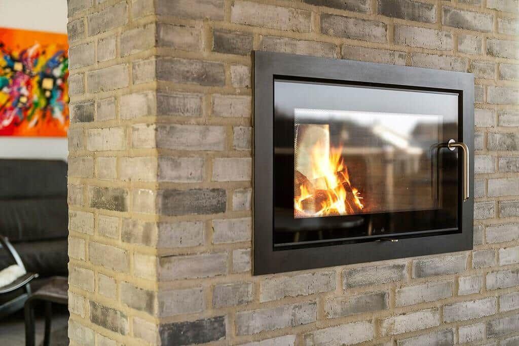 Ein Kamin mit brennendem Feuer in einer Ziegelwand. Im Hintergrund ist ein Sofa und eine Wanddekoration sichtbar.