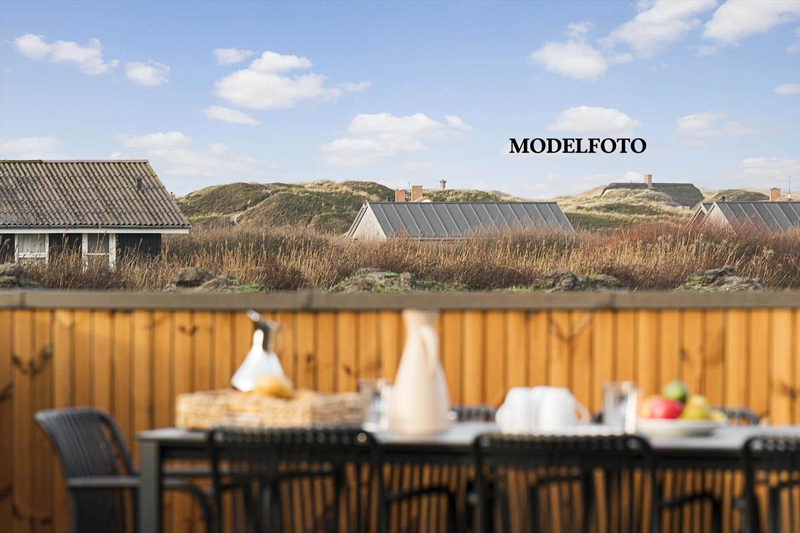 Terrasse mit Tisch und Stühlen. Hintergrund: Dünengrund mit Häusern und blauem Himmel.