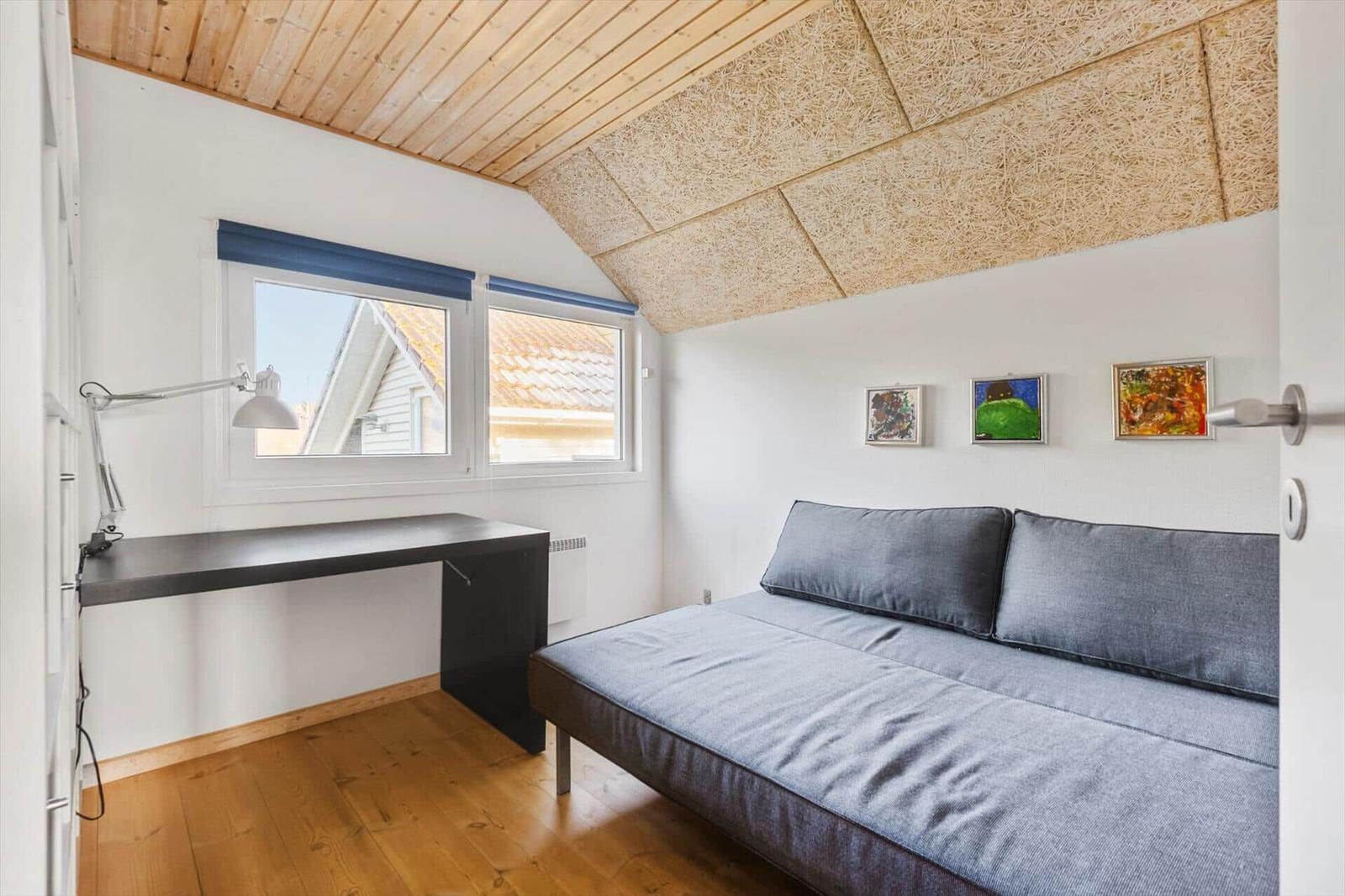 Zimmer mit Schlafcouch, Schreibtisch und Fenstern. Holzdecke und Wandbilder.