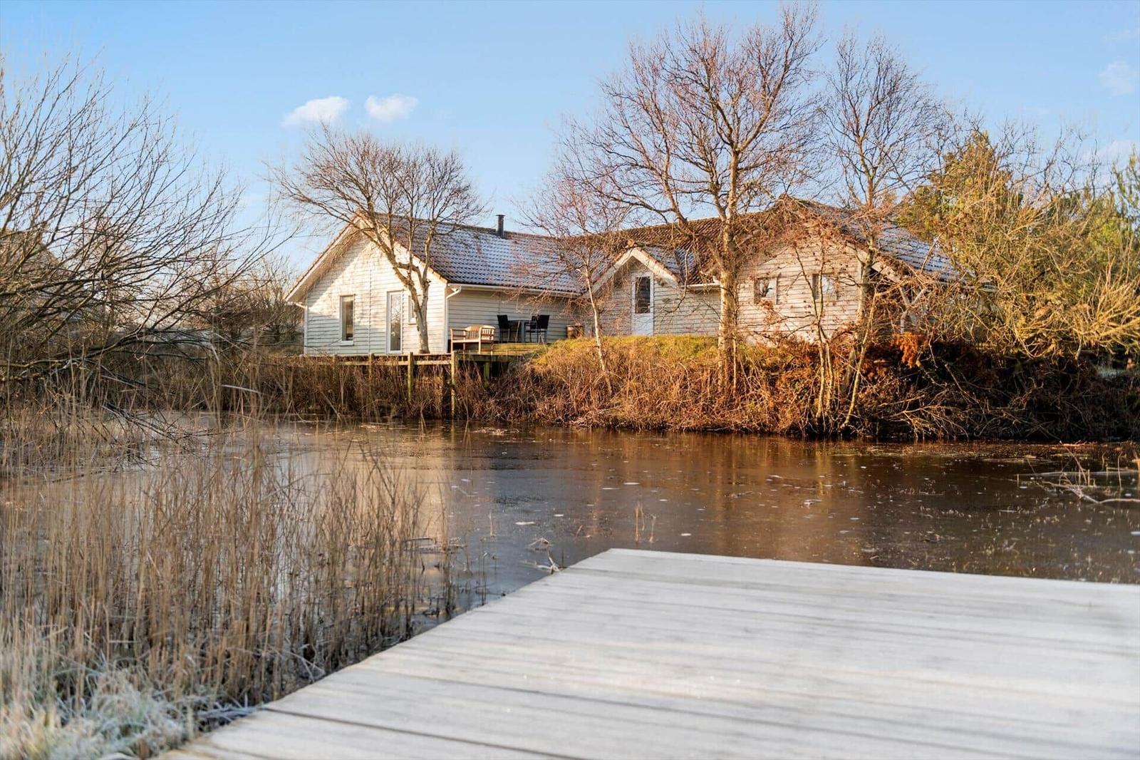 Haus am Wasser mit Holzsteg und Terrasse. Bäume und Schilf umgeben die ruhige Umgebung.