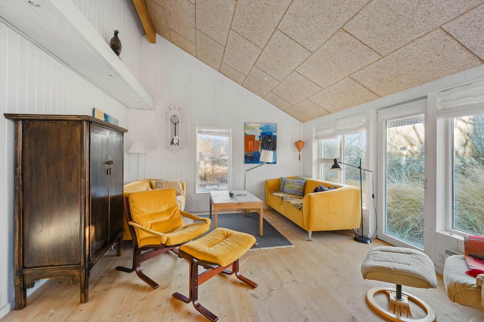 Wohnzimmer mit gelben Sitzmöbeln, Holzboden und großen Fenstern. Hölzerner Schrank und Wanduhr sichtbar.