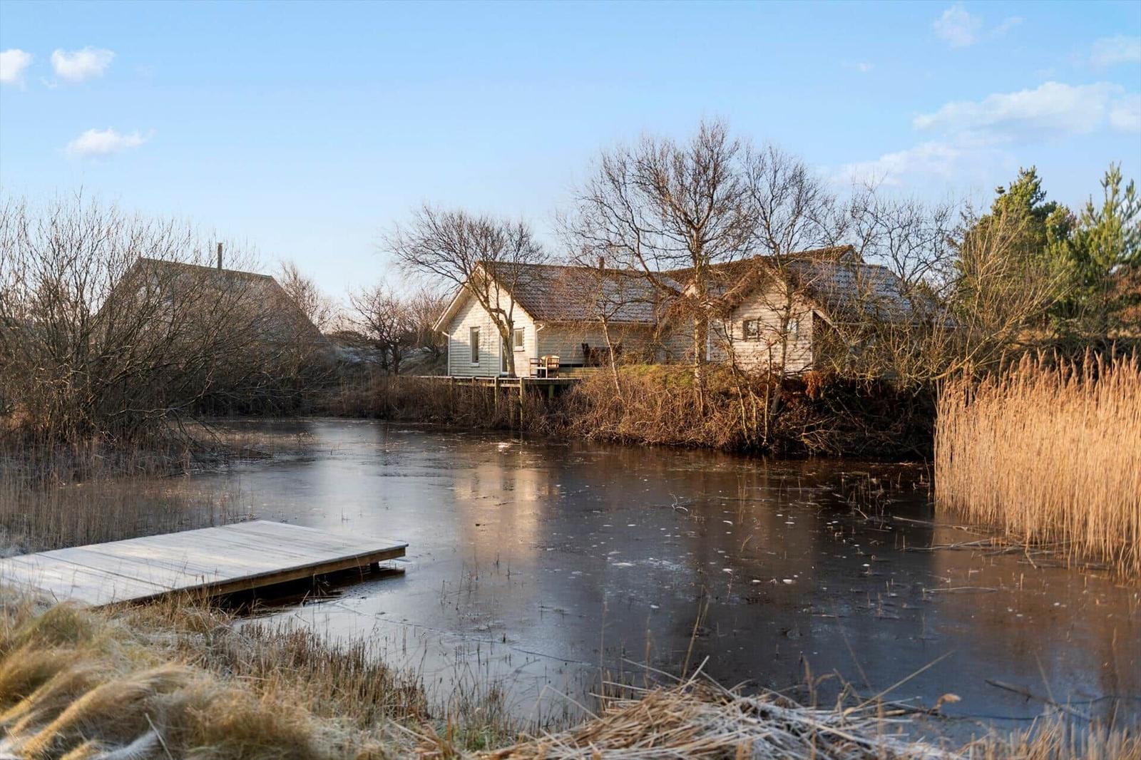 Haus am Ufer eines teilweise zugefrorenen Sees mit hölzerner Plattform und Ufervegetation.