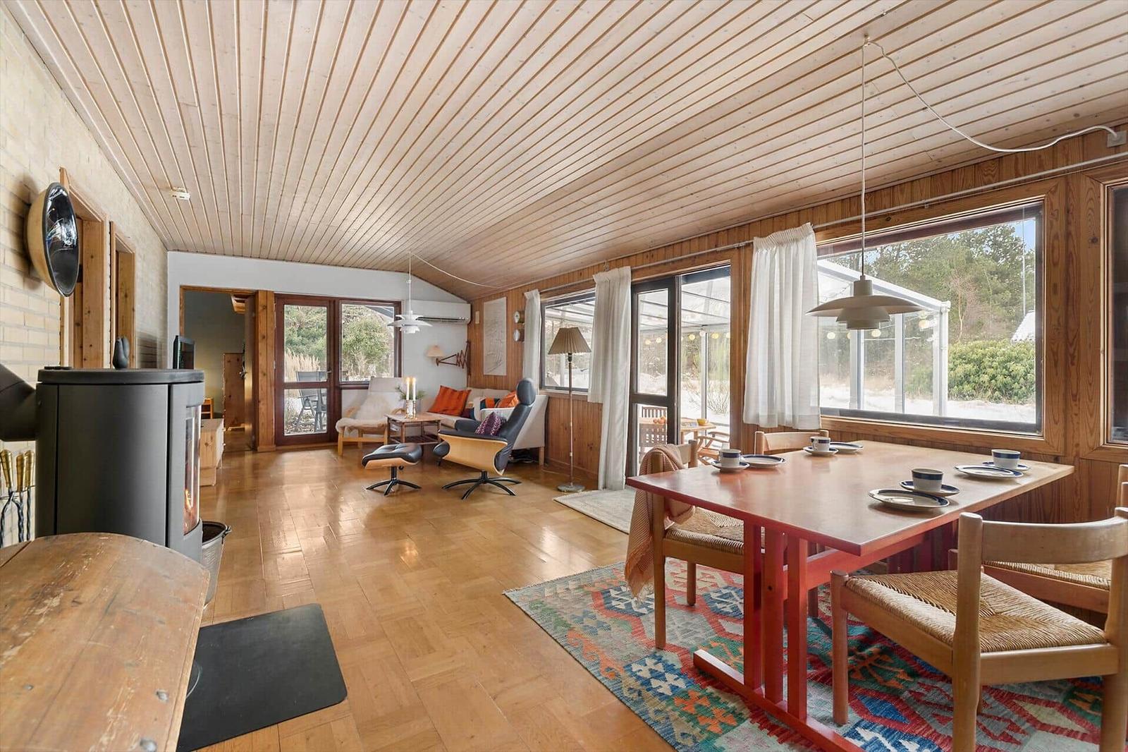 A room with dining area, living area, and wood ceiling. It has large windows and a fireplace.