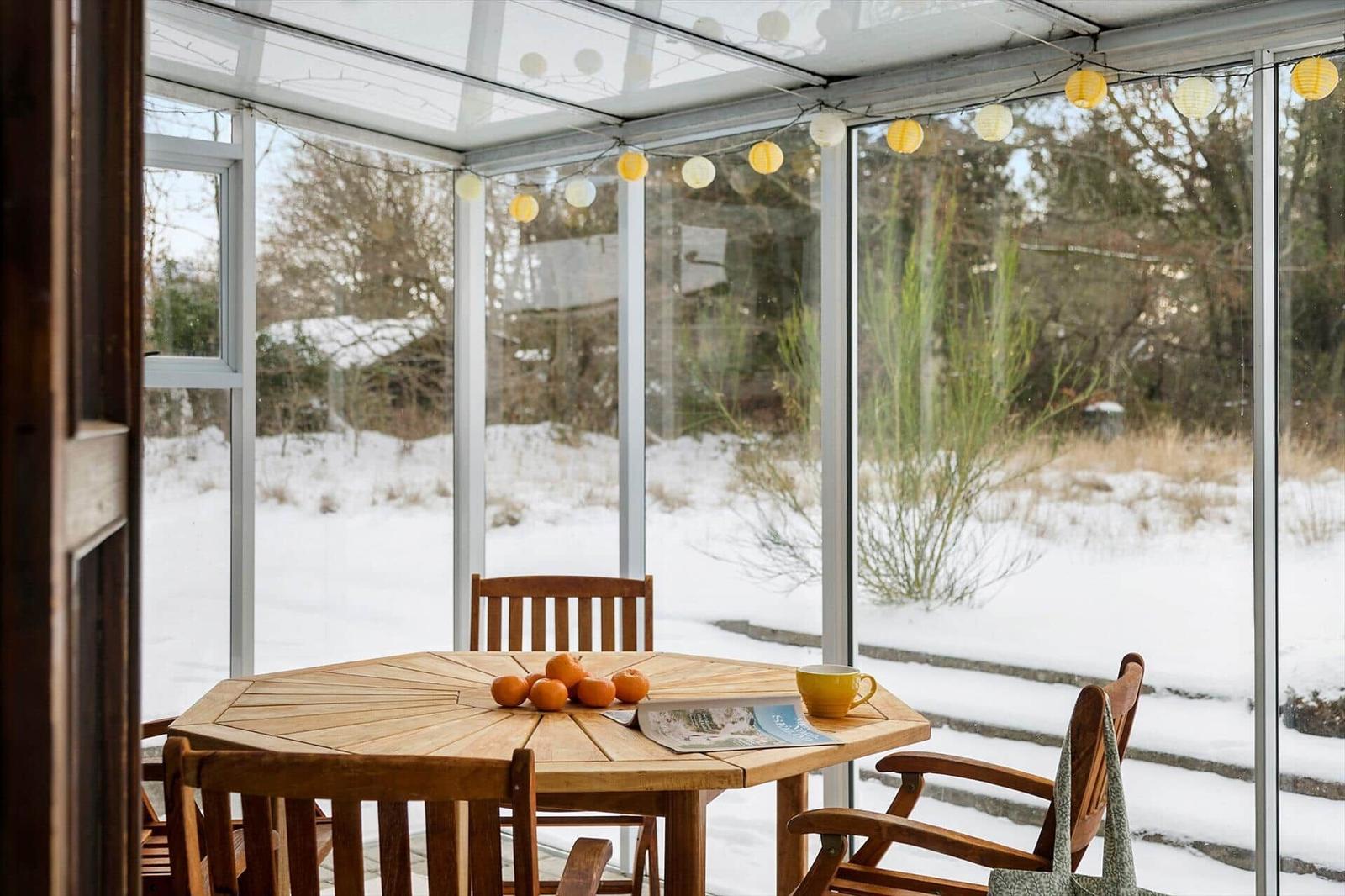 A wooden table with chairs, oranges, and a cup under a glass roof with string lights.
