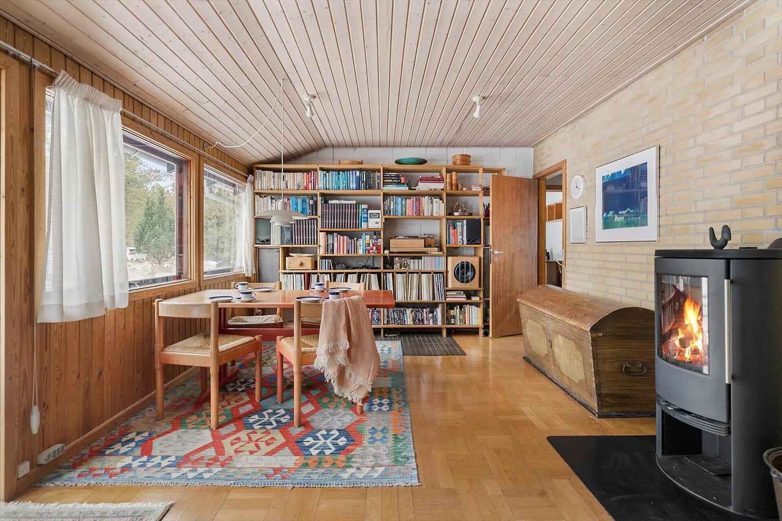 Dining area with wooden table, bookshelf, and wood-burning stove. Wooden walls and parquet floor.