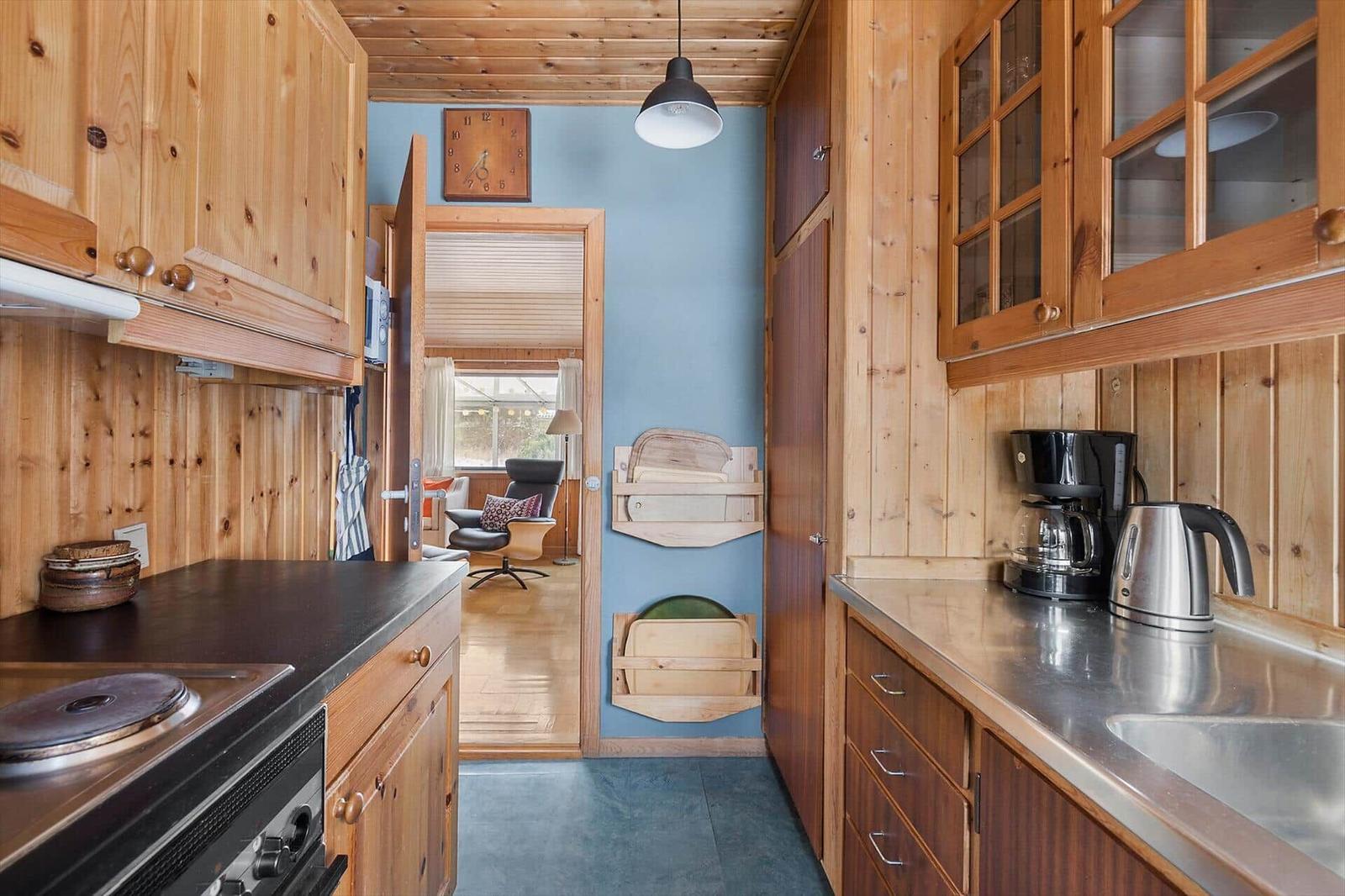 Kitchen with wooden furniture, coffee maker, and kettle. Corridor to living room with chair.