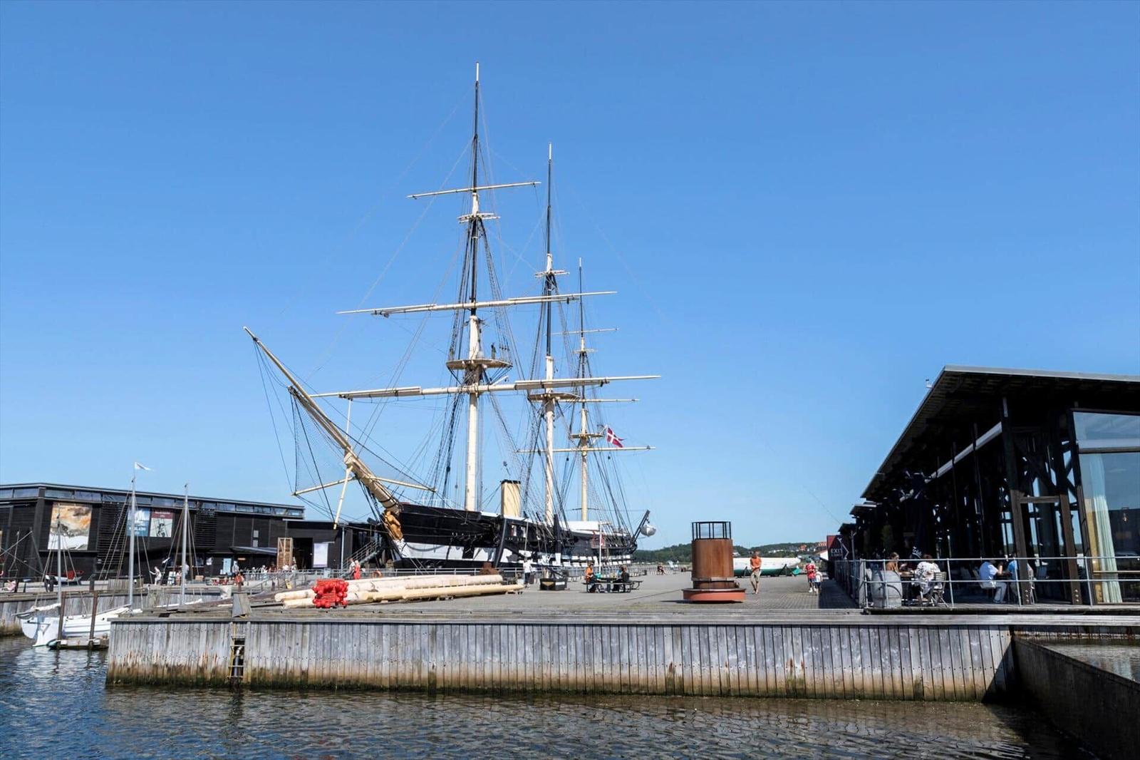 Historic sailing ship at the harbor with modern buildings and visitors.