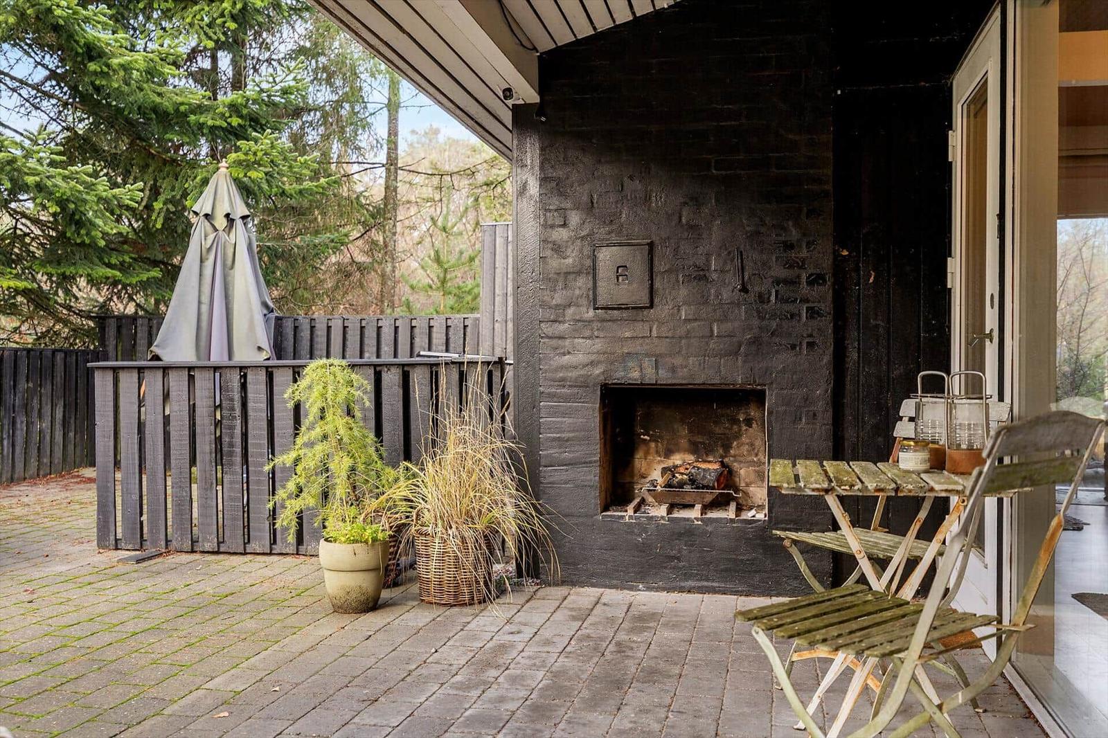 Outdoor area with stone fireplace, table, chairs, and plants. Background: wooden fence and trees.