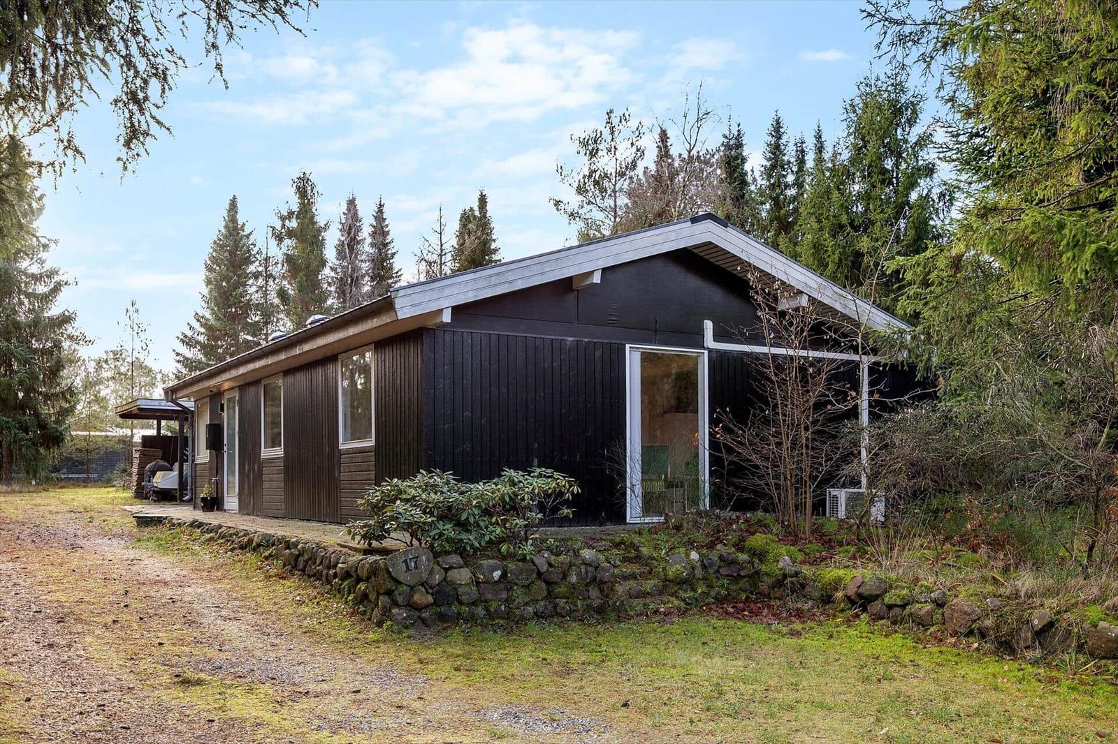 Black wooden house with glass front, surrounded by trees and stone wall.