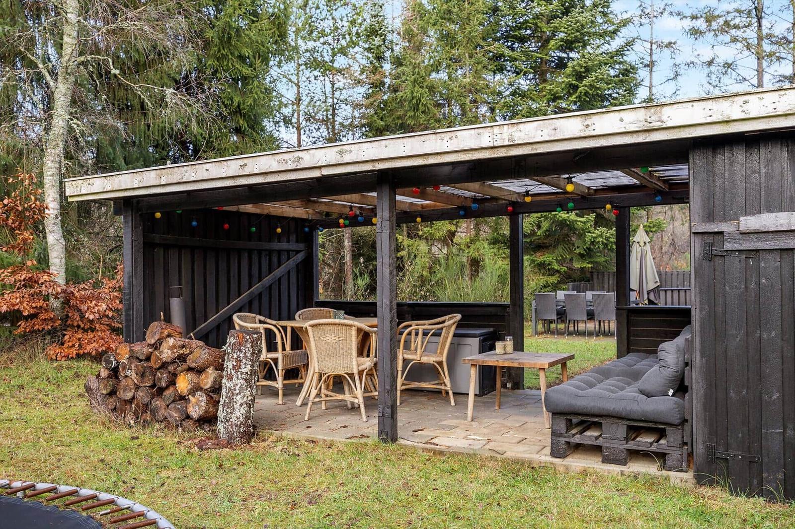 Outdoor area with seating, table and chairs under roof. Woodpile and grill area visible.