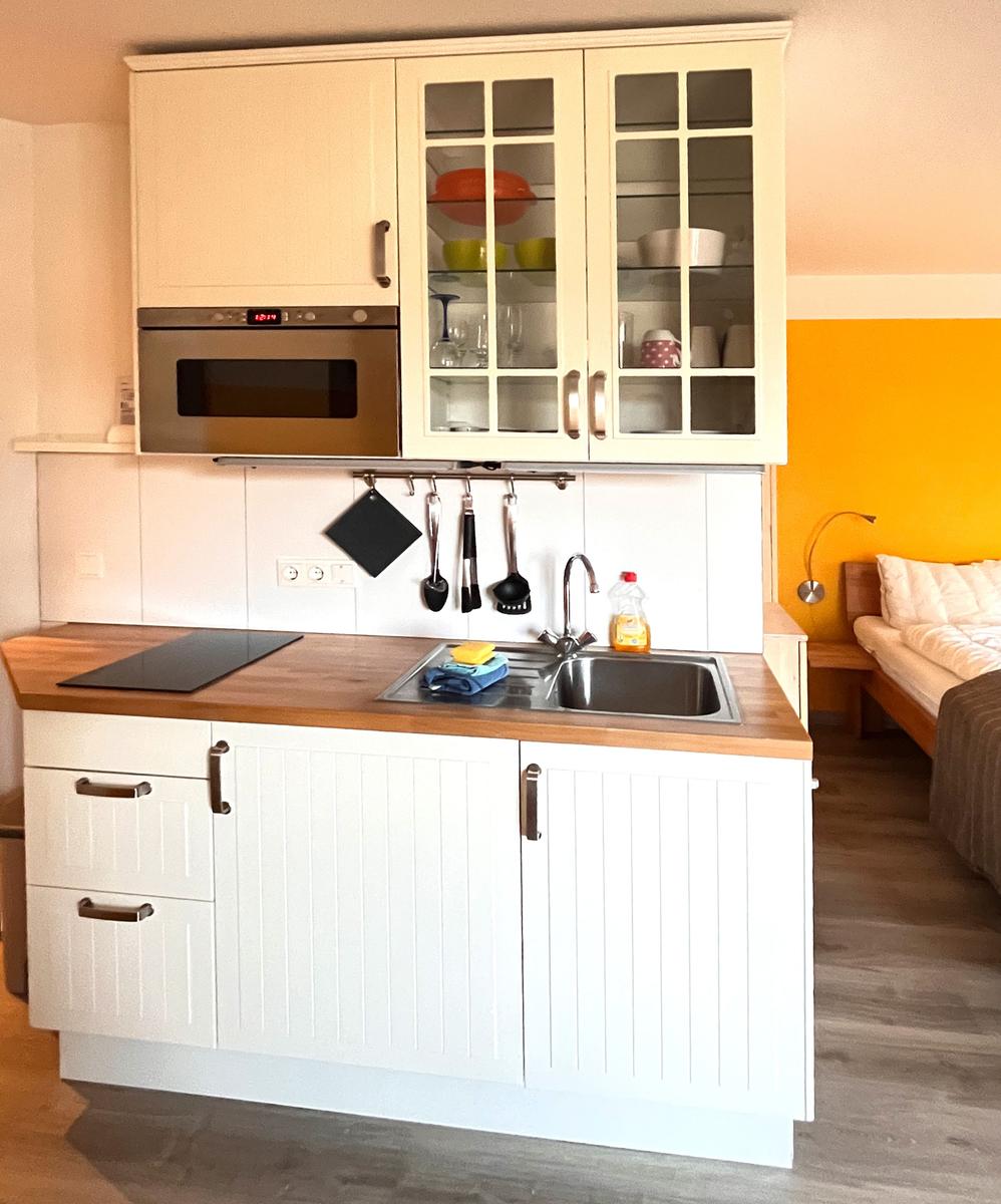 Küche mit weißen Schränken, Edelstahlspüle und integriertem Ofen. Im Hintergrund ein Bett.