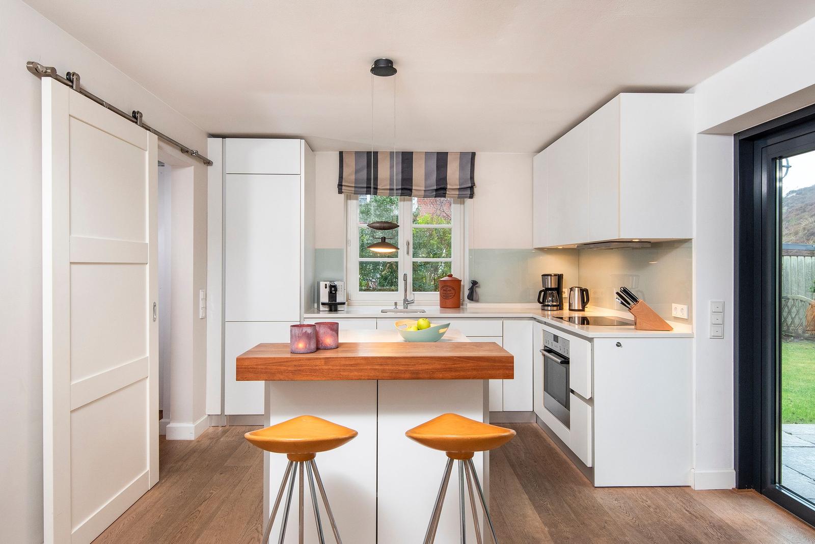 Küche mit Kücheninsel, weißen Schränken und Holzboden. Fenster und Glastür bieten Blick auf Garten.