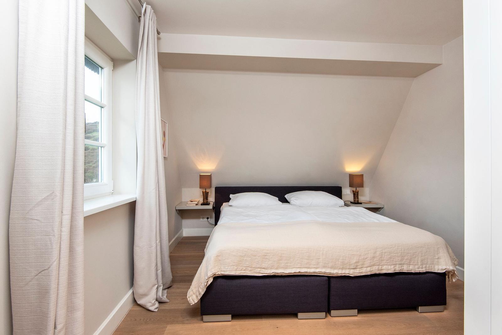 Doppelbett mit weißen Kissen und Bettwäsche. Fenster mit weißen Vorhängen. Holzboden und Nachttische mit Lampen.