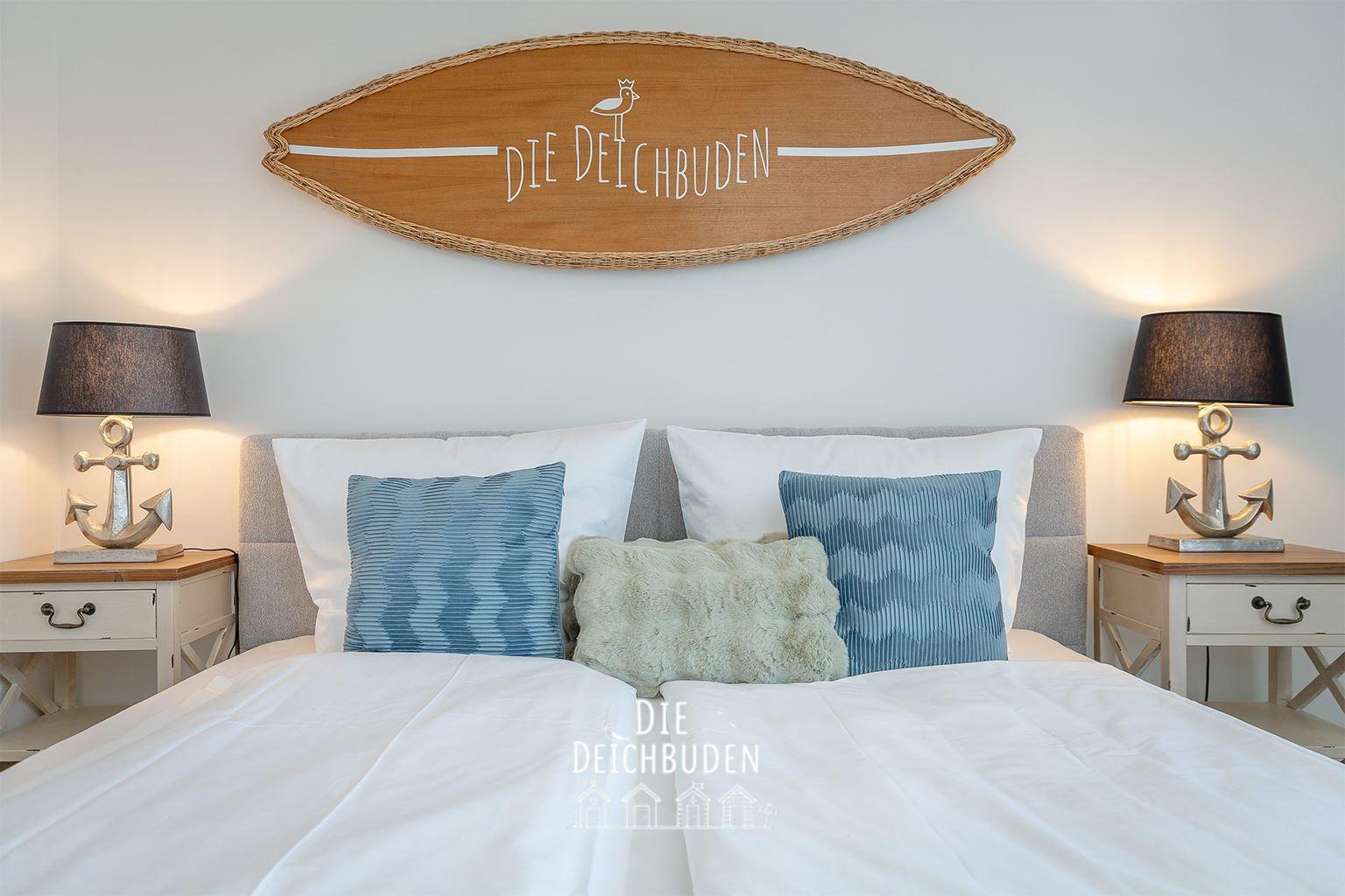 Schlafzimmer mit Bett, Kissen und Nachttischen mit Ankerlampen. Wanddekoration mit Surfbrett.