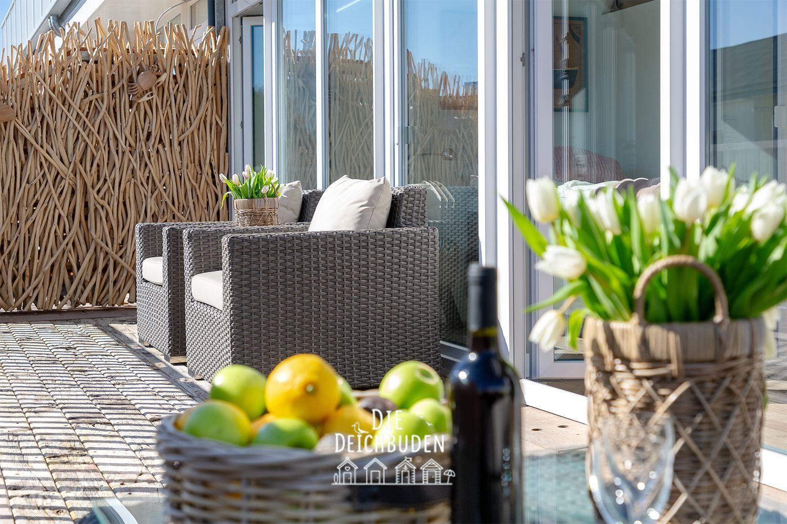 Terrasse mit Sitzgruppe, Blumen und Obst. Hintergrund: Glasfront und Holz-Zaun.