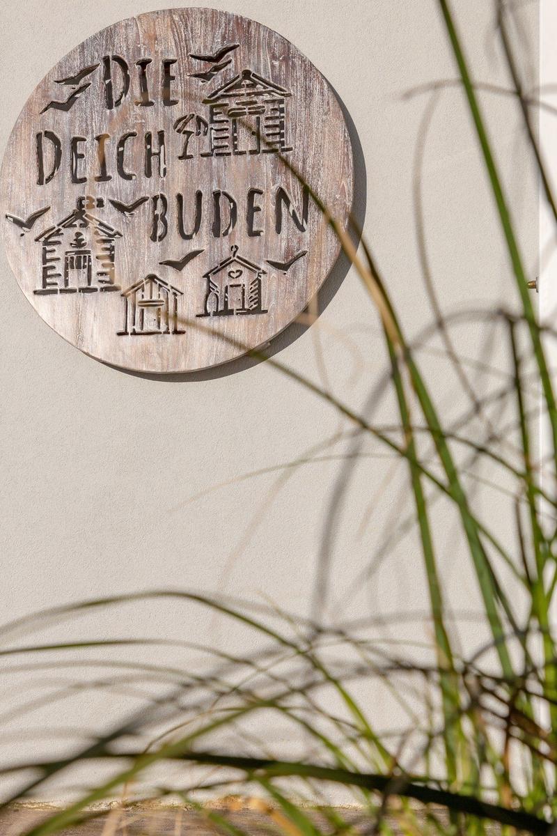 Rundes Holzschild mit Schriftzug und Hausmotiven an Wand, Gräser im Vordergrund.