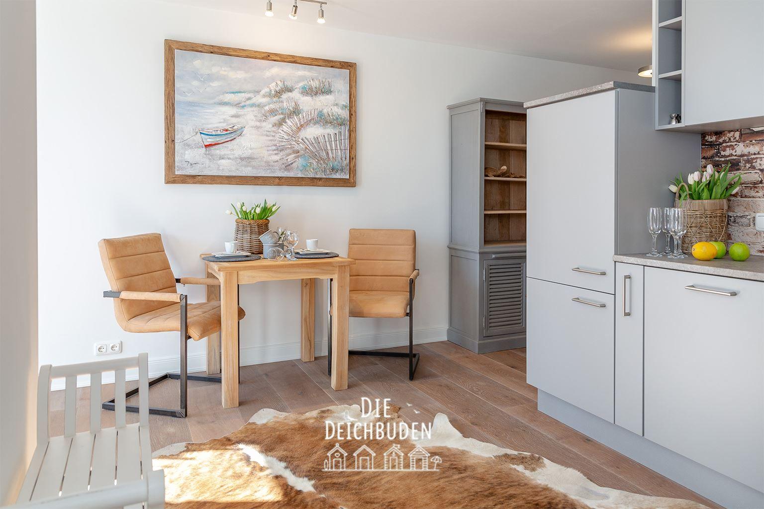 Essbereich mit Tisch, zwei Stühlen und Küchenzeile. Wandbild zeigt Strandmotiv.