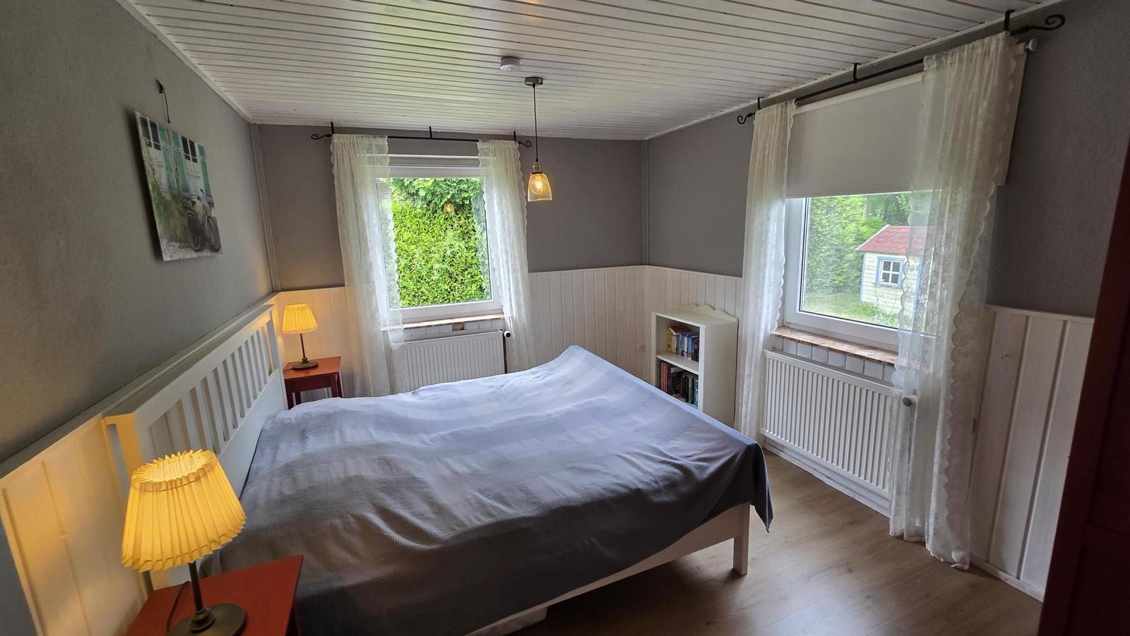 Schlafzimmer mit Bett, zwei Fenstern und Nachttischen.