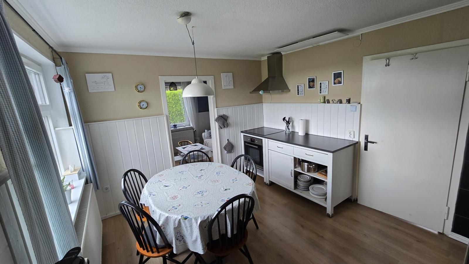 Küche mit Esstisch, Herd und Fenster. Weiße Schränke und Holzboden.