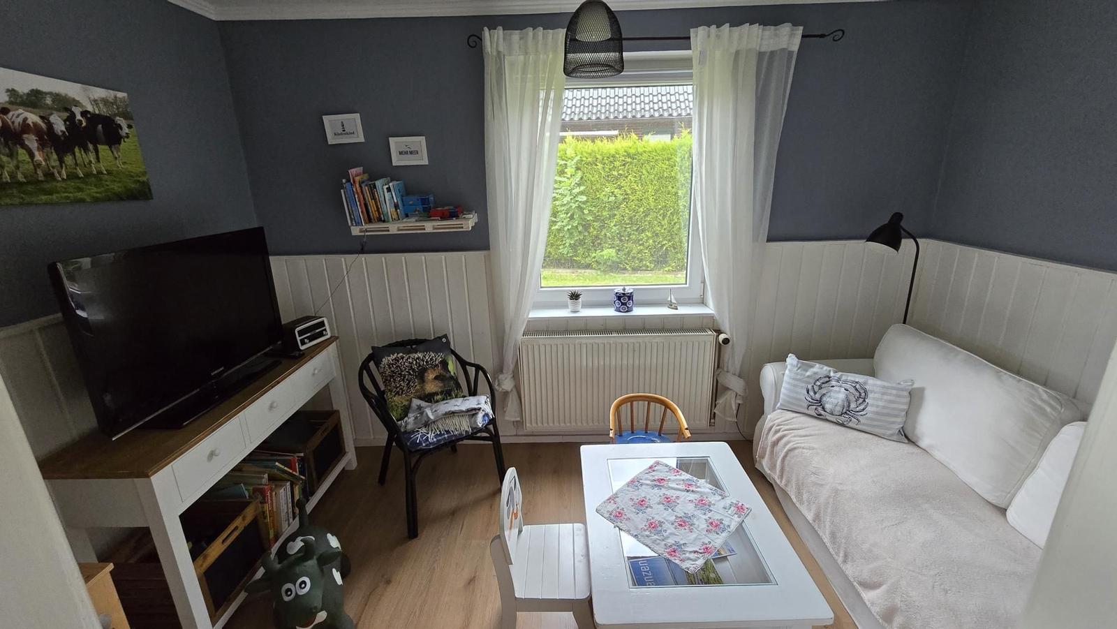 Ein Wohnzimmer mit Sofa, TV, Tisch und Stühlen. Fenster mit Vorhängen und Wandbild.