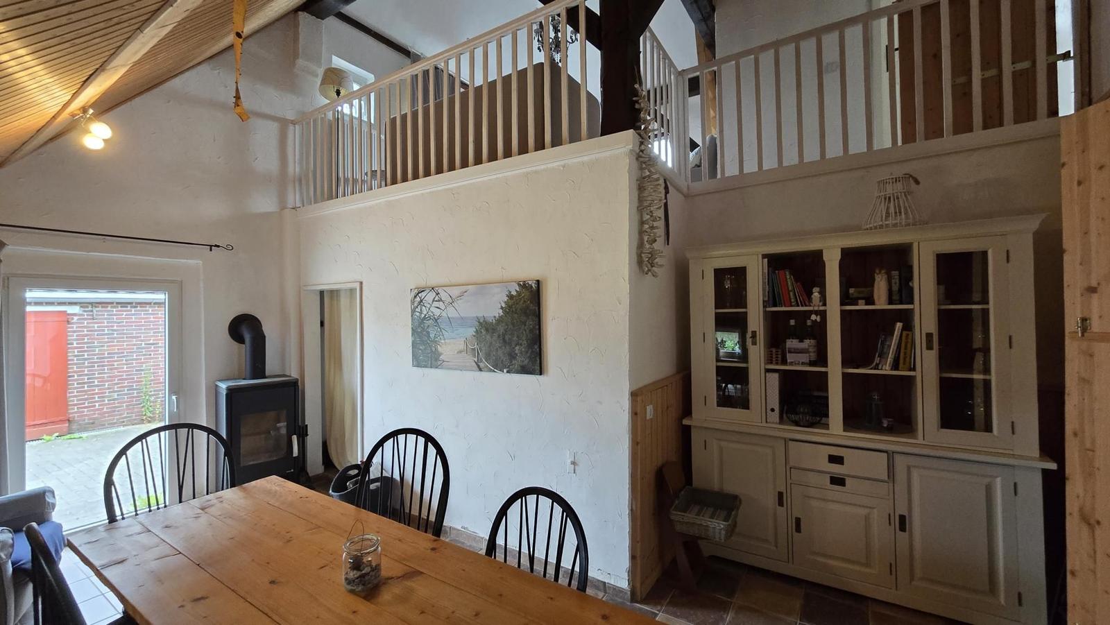 Essbereich mit Holztisch, schwarzen Stühlen und Kaminofen. Obergeschoss mit Balkon.